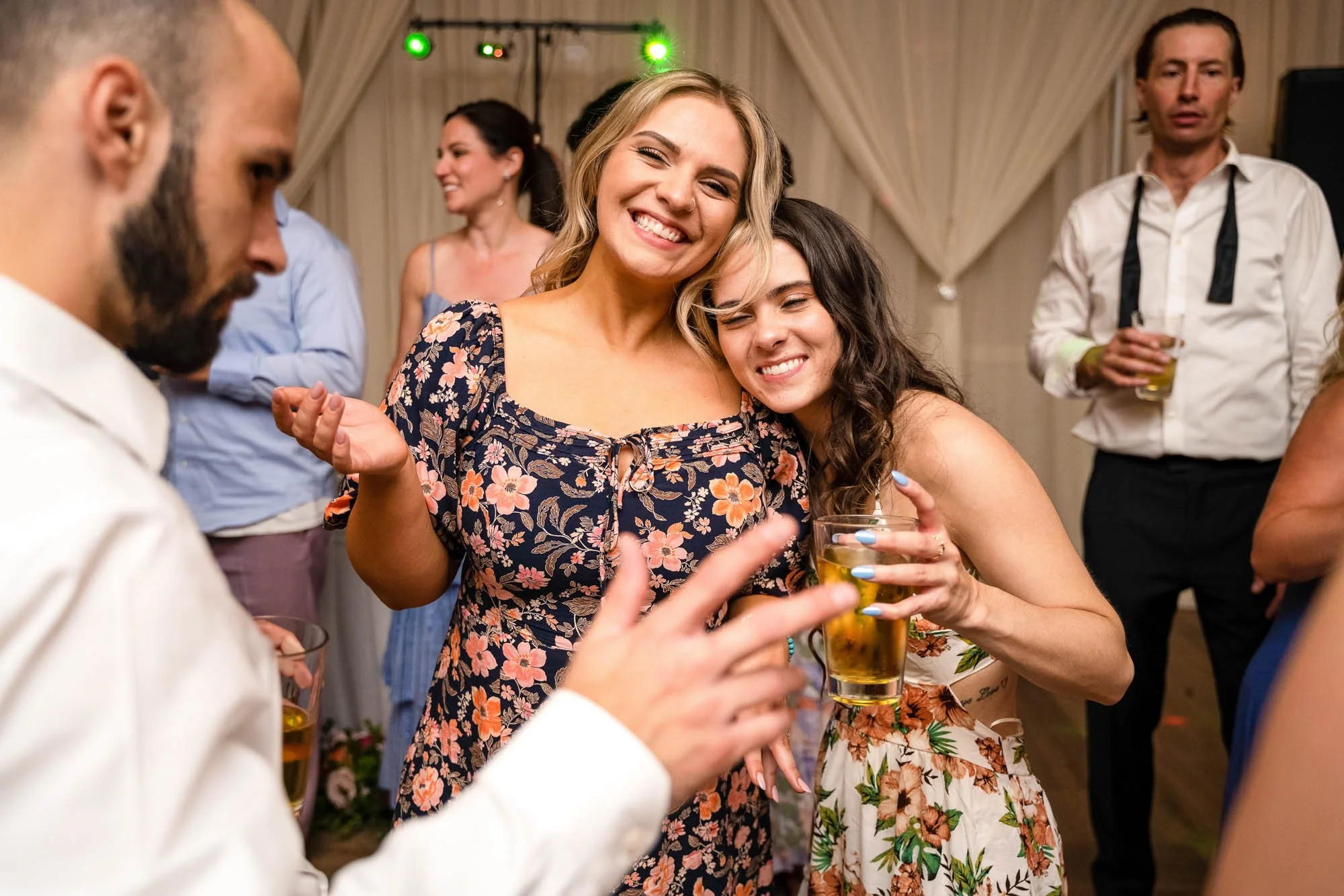 Wedding photograph at Abernethy Center Veiled Garden and Ballroom of Two women hugging and smiling at a social gathering, holding drinks, with other guests in the background.