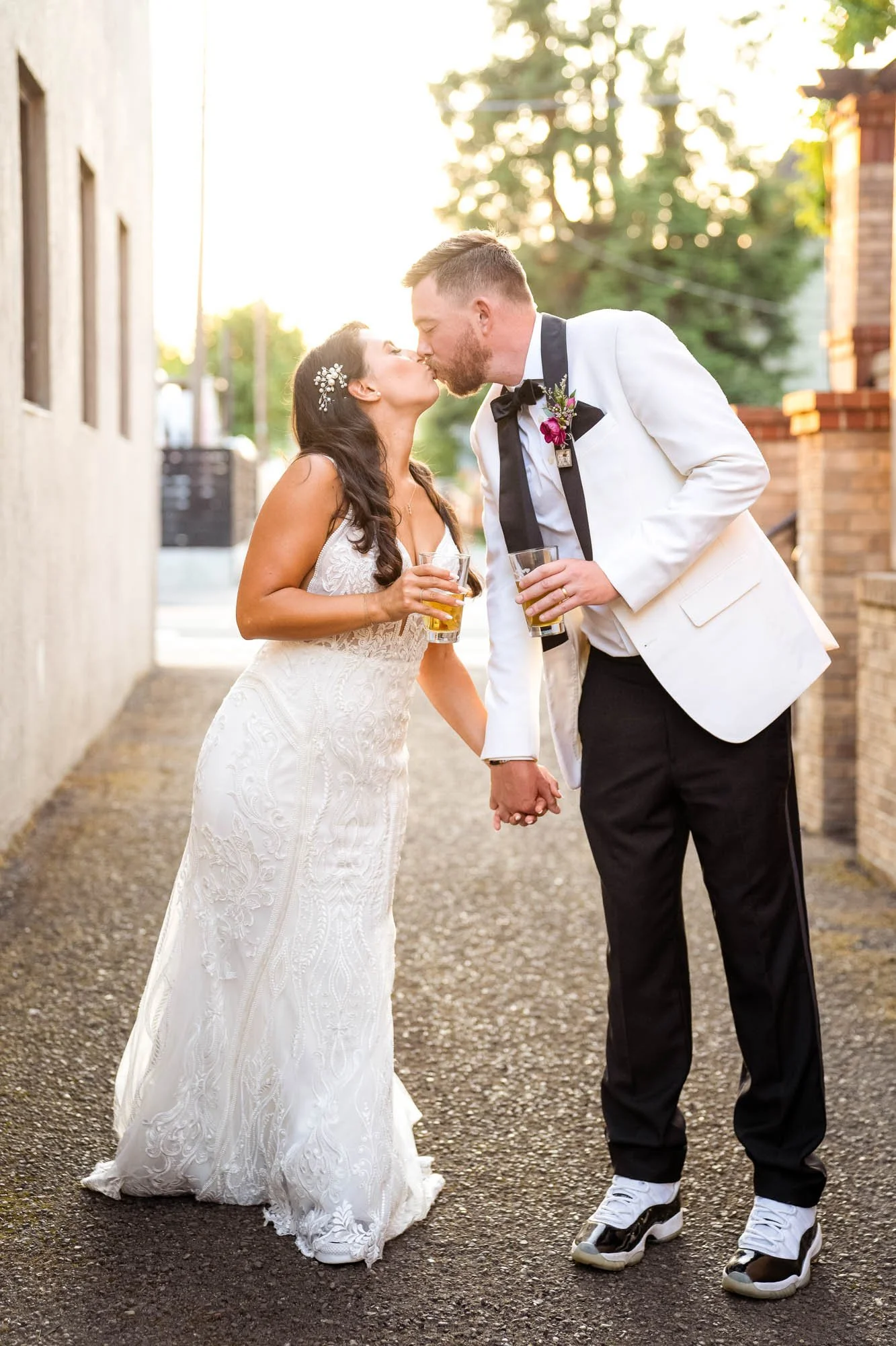 A bride and groom share a kiss while holding hands and drinking beer outdoors during sunset at Abernethy Center Veiled Garden and Ballroom.