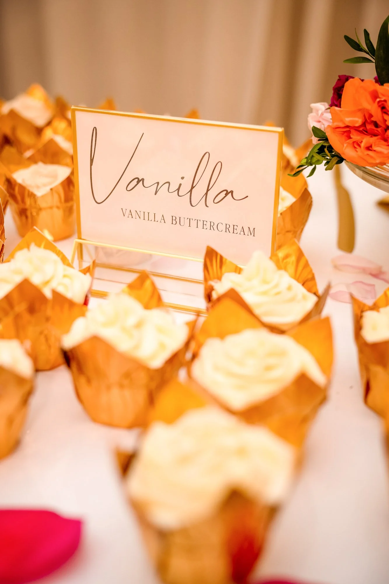 Cupcakes with vanilla frosting on a white table, decorated with a pink sign labeled 'Vanilla' and 'Vanilla Buttercream', surrounded by pink and orange flowers.