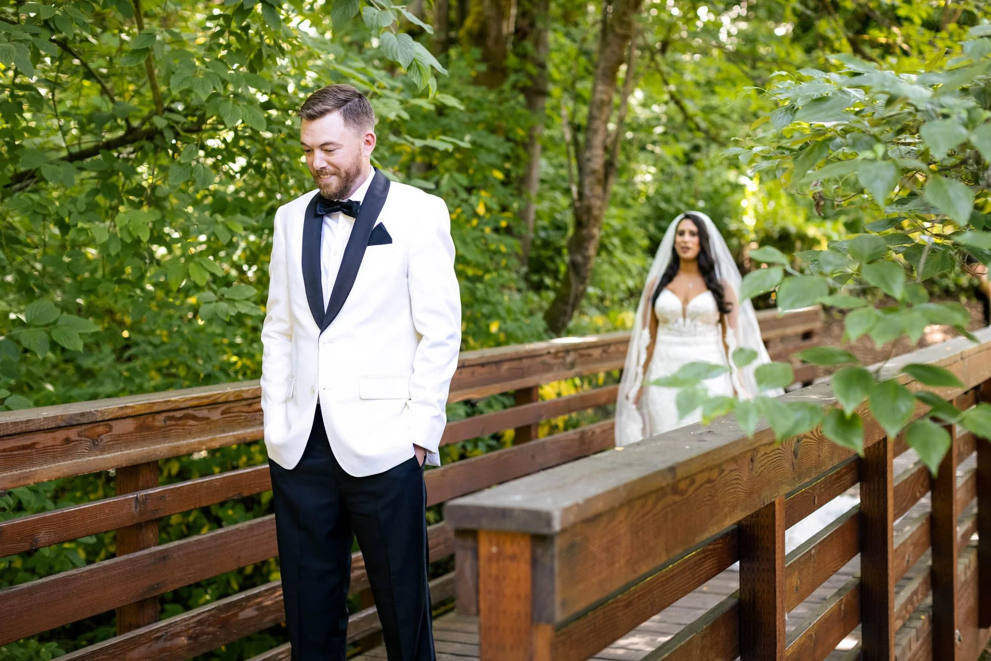 A groom in a white tuxedo jacket with black lapels and bow tie, smiling and looking down with his hands in his pockets, standing on a wooden bridge in a lush green outdoor setting. In the background, a bride in a white wedding dress and veil is walki