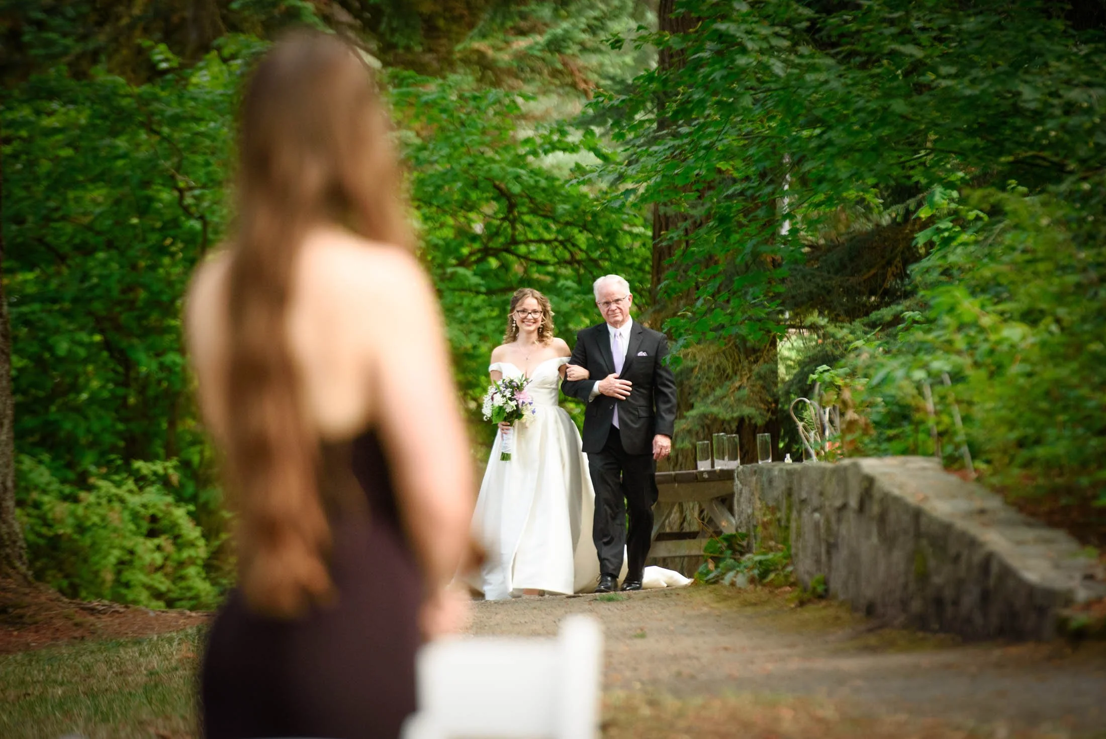 Wedding photo at Hoyt Arboretum of A bride walking down the aisle with her father at an outdoor wedding ceremony surrounded by trees.