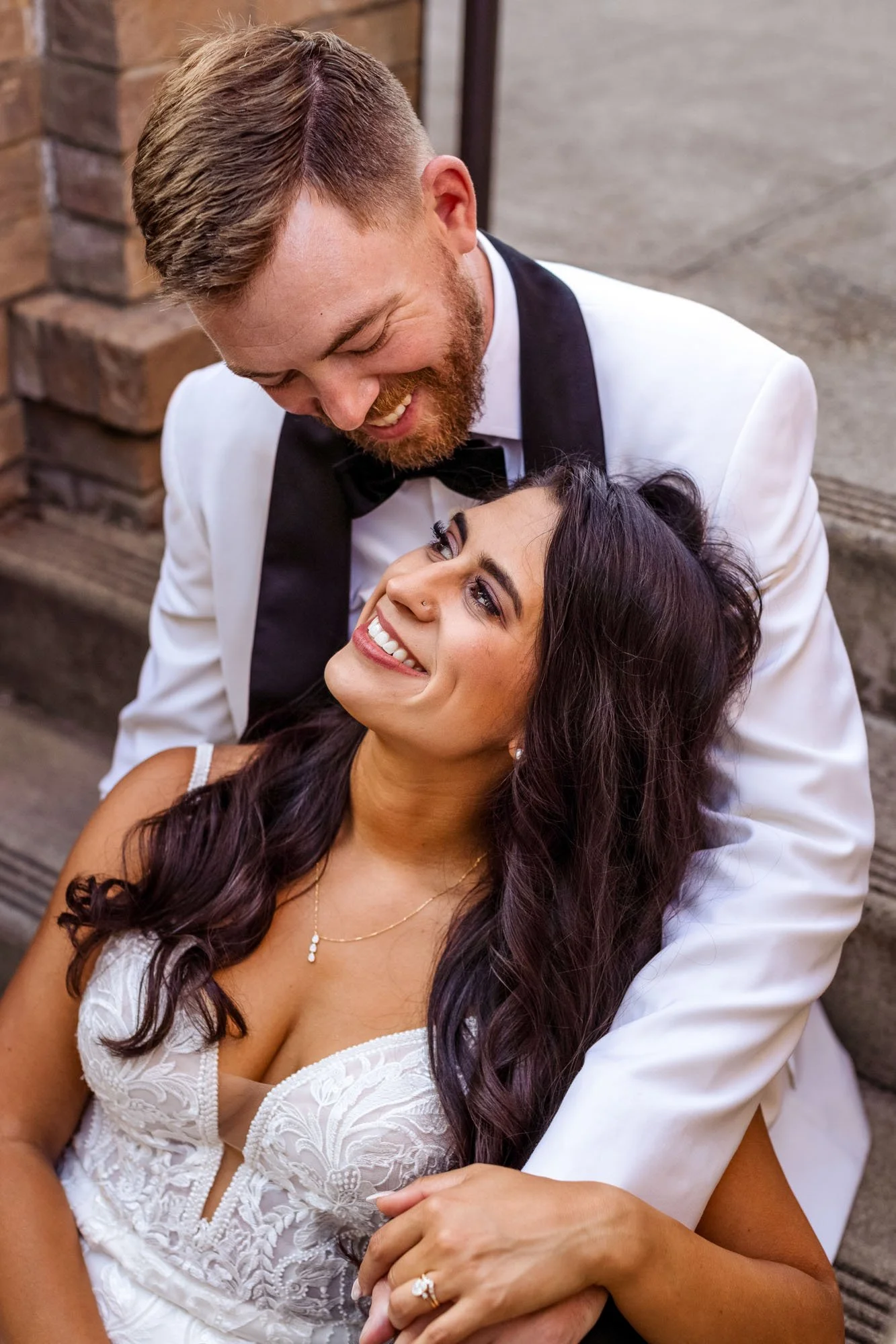 Wedding photograph at Abernethy Center Veiled Garden and Ballroom of A happy couple dressed in wedding attire, with the man in a white tuxedo and the woman in a white wedding dress, sitting on wooden stairs, embracing and smiling at each other.