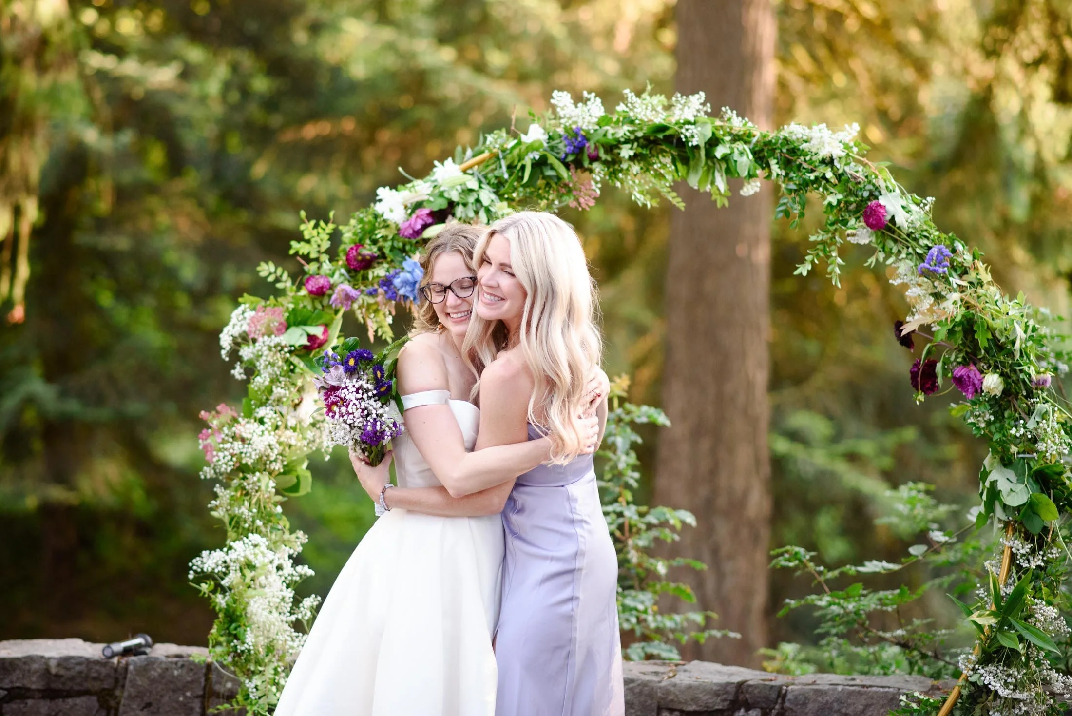 Wedding photograph at Portland's Hoyt Arboretum of Two women, one in a white dress and the other in a light purple dress, hugging and smiling in front of a floral arch outdoors.