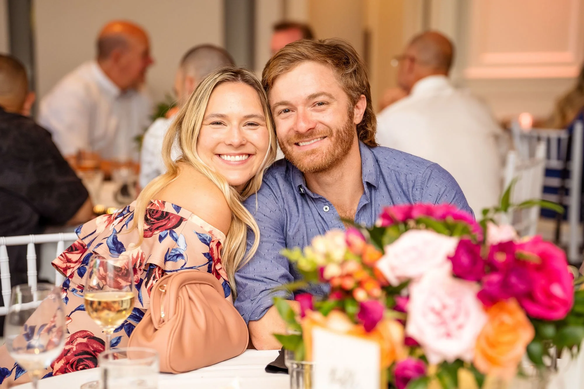 Wedding photo at Abernethy Center's Abernethy Chapel of a smiling young couple sitting at a table during a social event or wedding reception, with colorful flower arrangements and glasses of wine in the foreground.