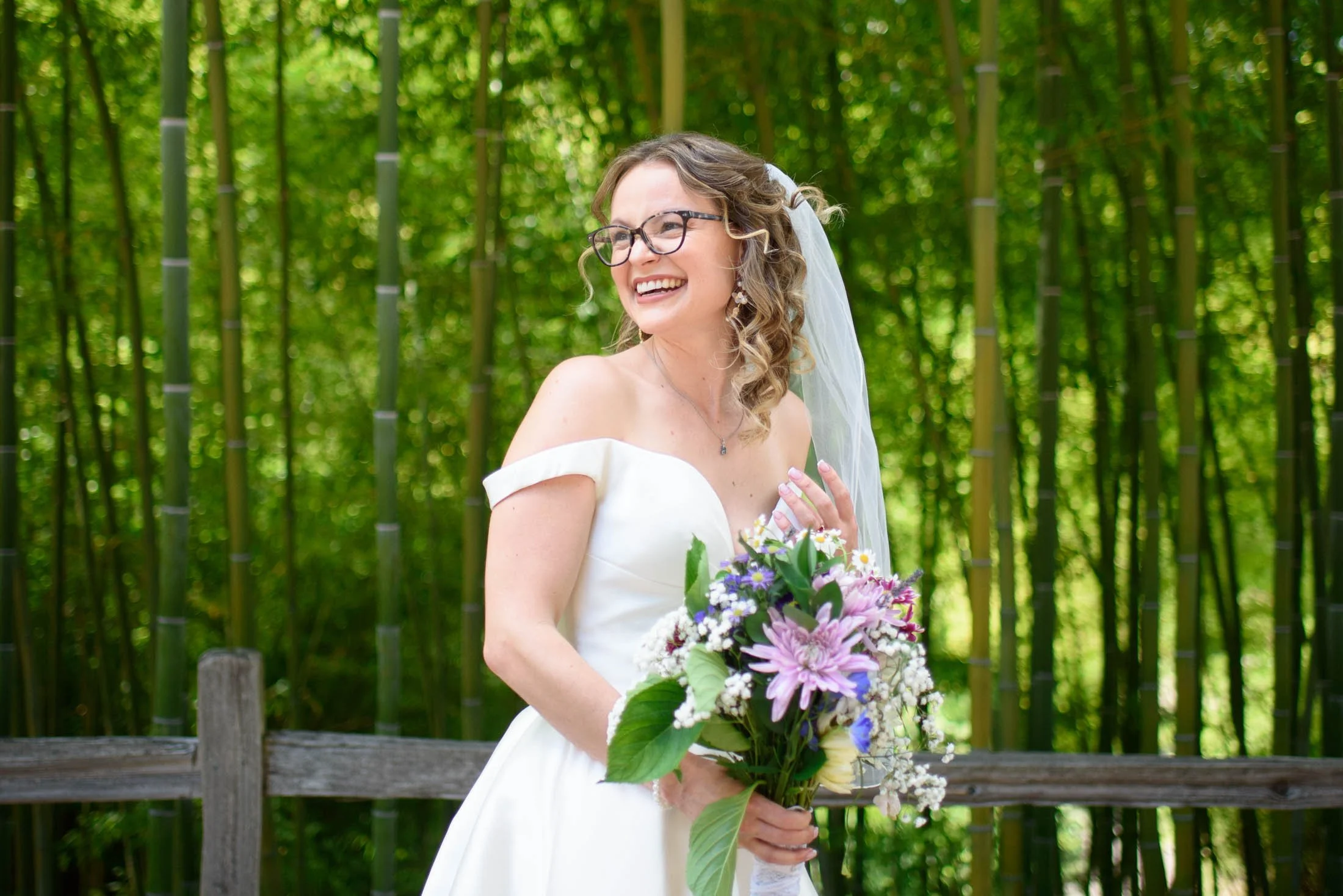 Wedding photo at Hoyt Arboretum of A bride in a white off-shoulder wedding dress holding a bouquet of flowers, smiling outdoors in a green forest with bamboo trees in the background.