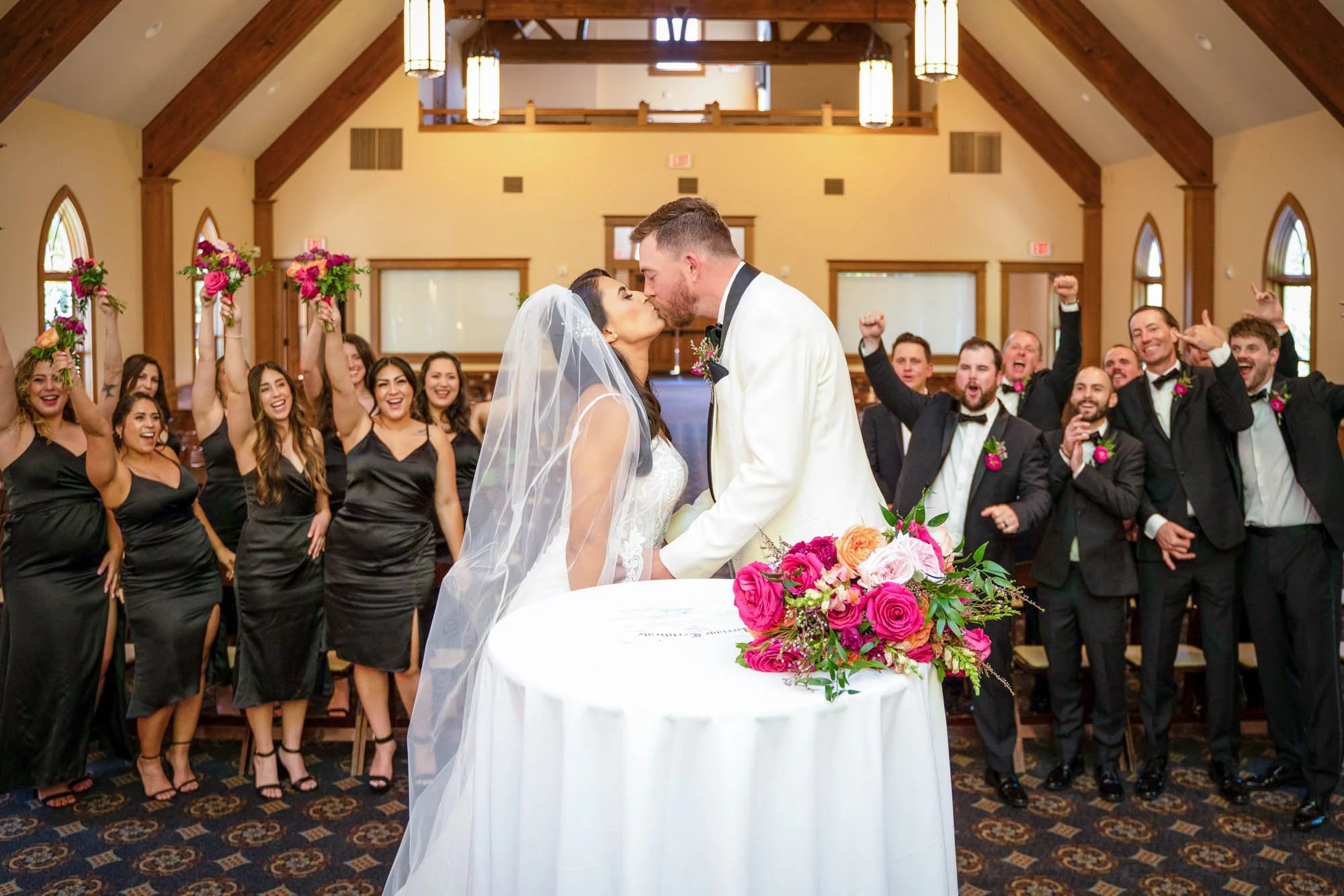Wedding photography at Abernethy Center's Chapel of  a bride and groom kissing, surrounded by bridesmaids and groomsmen in a church or chapel setting, with cheerful expressions and colorful floral arrangements.