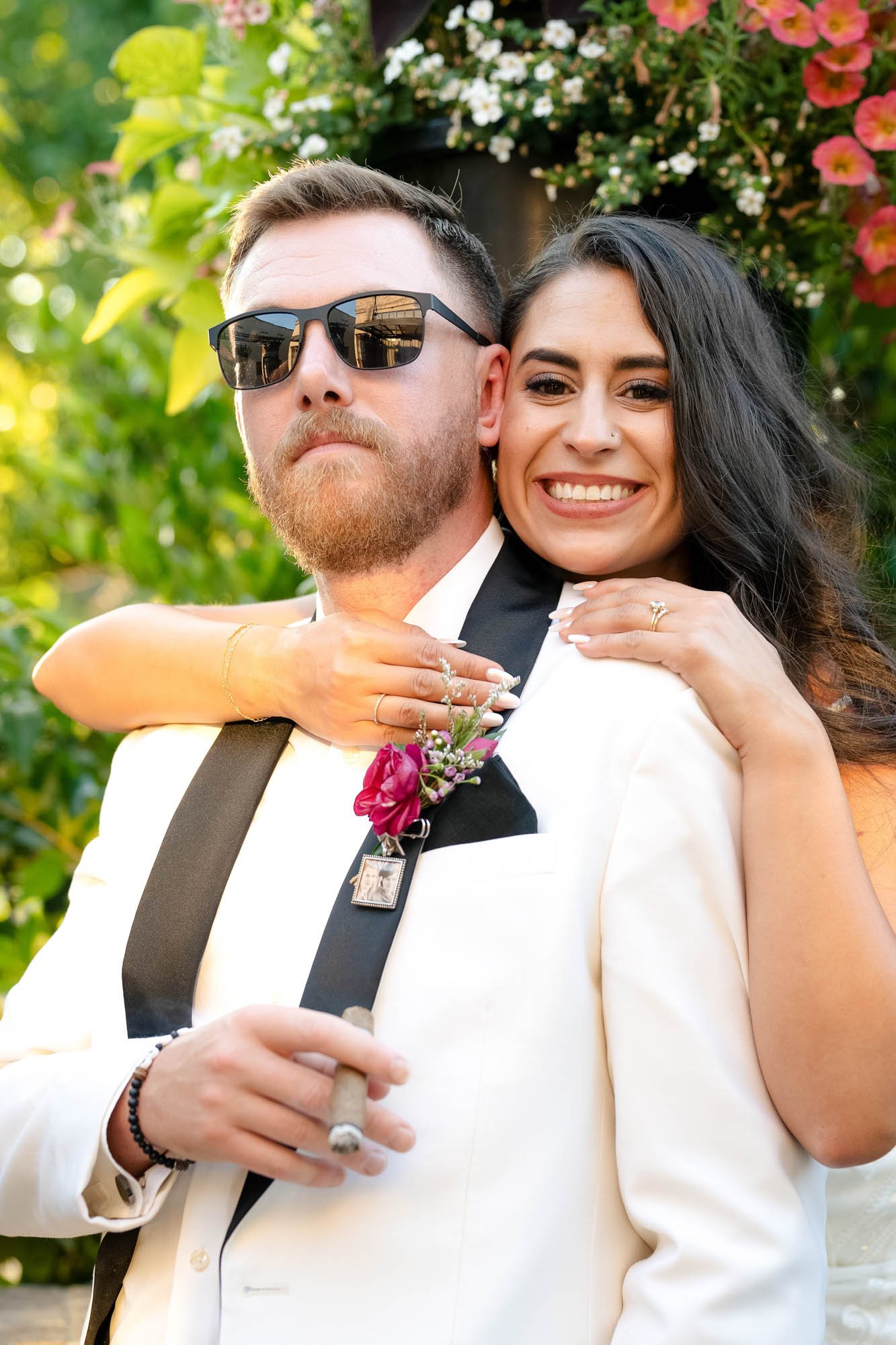 A smiling woman with dark hair and a nose piercing hugging a man in a white tuxedo with a boutonniere, sunglasses, and a cigar in an outdoor setting with green foliage and flowers at Abernethy Center Veiled Garden and Ballroom.