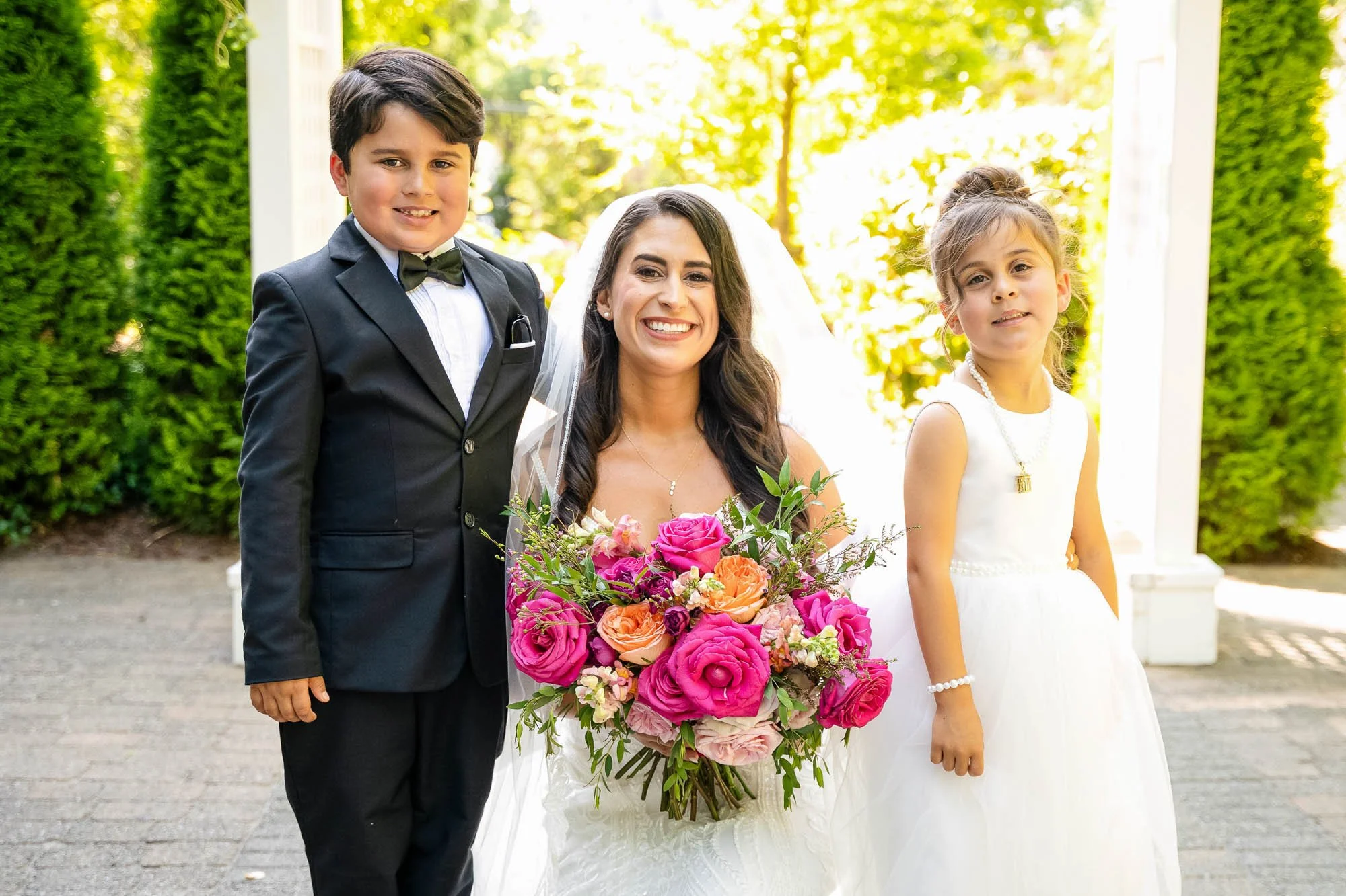 Smiling bride in white dress holding a colorful flower bouquet, standing between a young boy in a black suit and bow tie, and a young girl in a white dress with pearl necklace, outdoors at Abernethy Center.