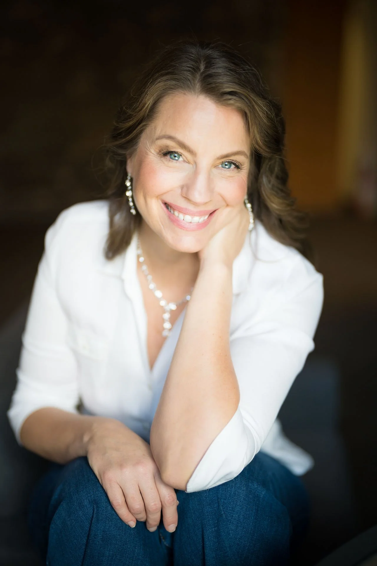 Professional Close-up portrait of a smiling woman with blue eyes, wearing pearl earrings, a pearl necklace, a white blouse, and blue jeans, sitting indoors with a blurred wooden background.