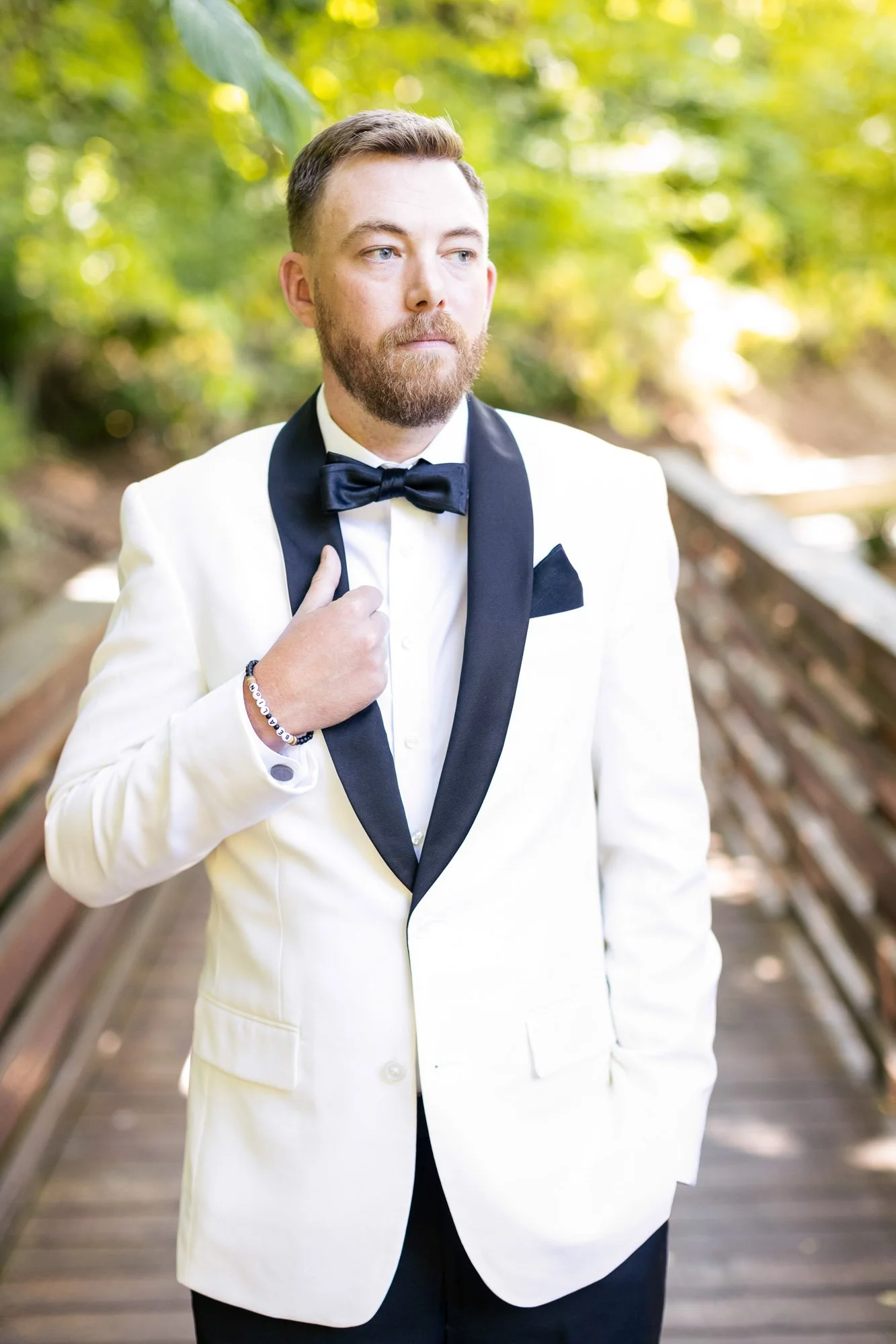 Wedding photo at Abernethy Center of A man in a white tuxedo jacket, black bow tie, and black pants standing outdoors on a wooden bridge, adjusting his tuxedo. The background features green trees and sunlight.