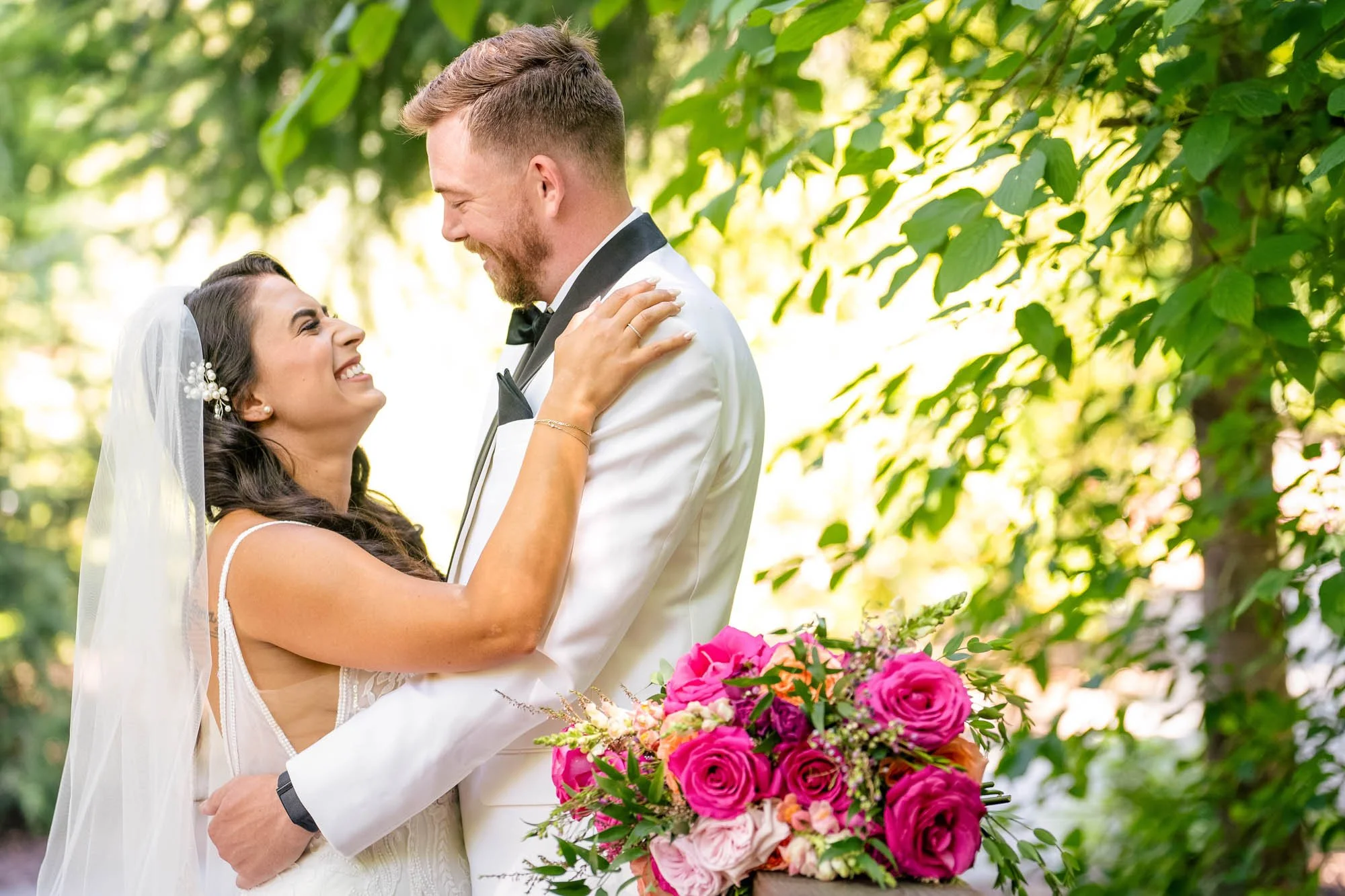 Wedding photo at Abernethy Center of A happy bride and groom embracing outdoors, with the bride holding a bouquet of pink roses and greenery, surrounded by lush green foliage.