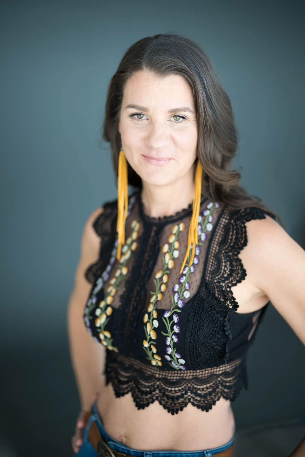 Headshot of A woman with dark hair wearing a black embroidered lace top with yellow earrings.