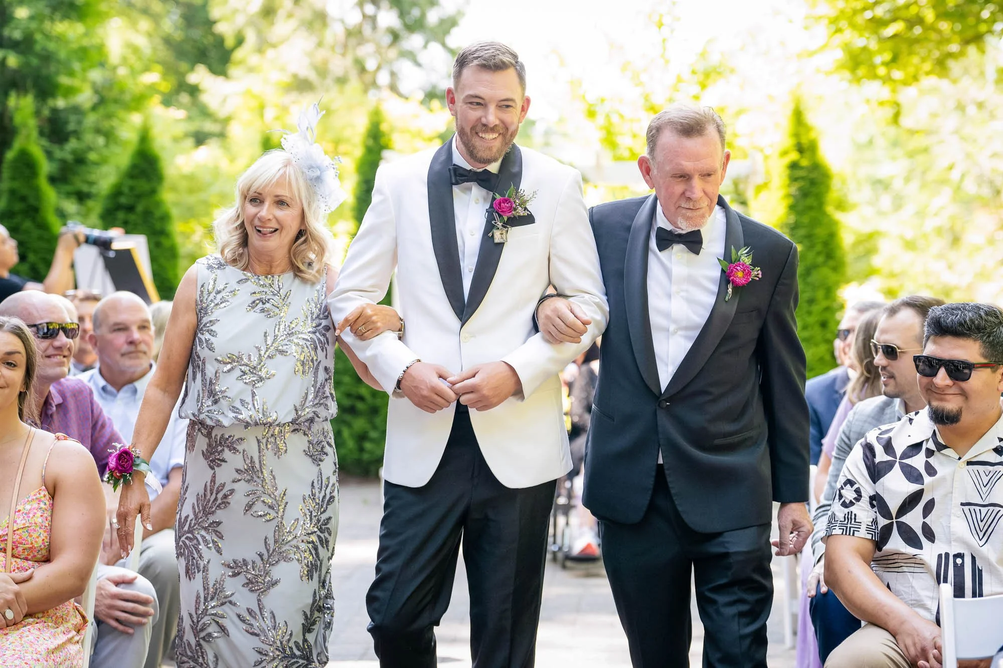 Wedding photography at Abernethy Center of  A groom in a white tuxedo with black lapel, walking arm-in-arm with his parents at an outdoor wedding ceremony. 