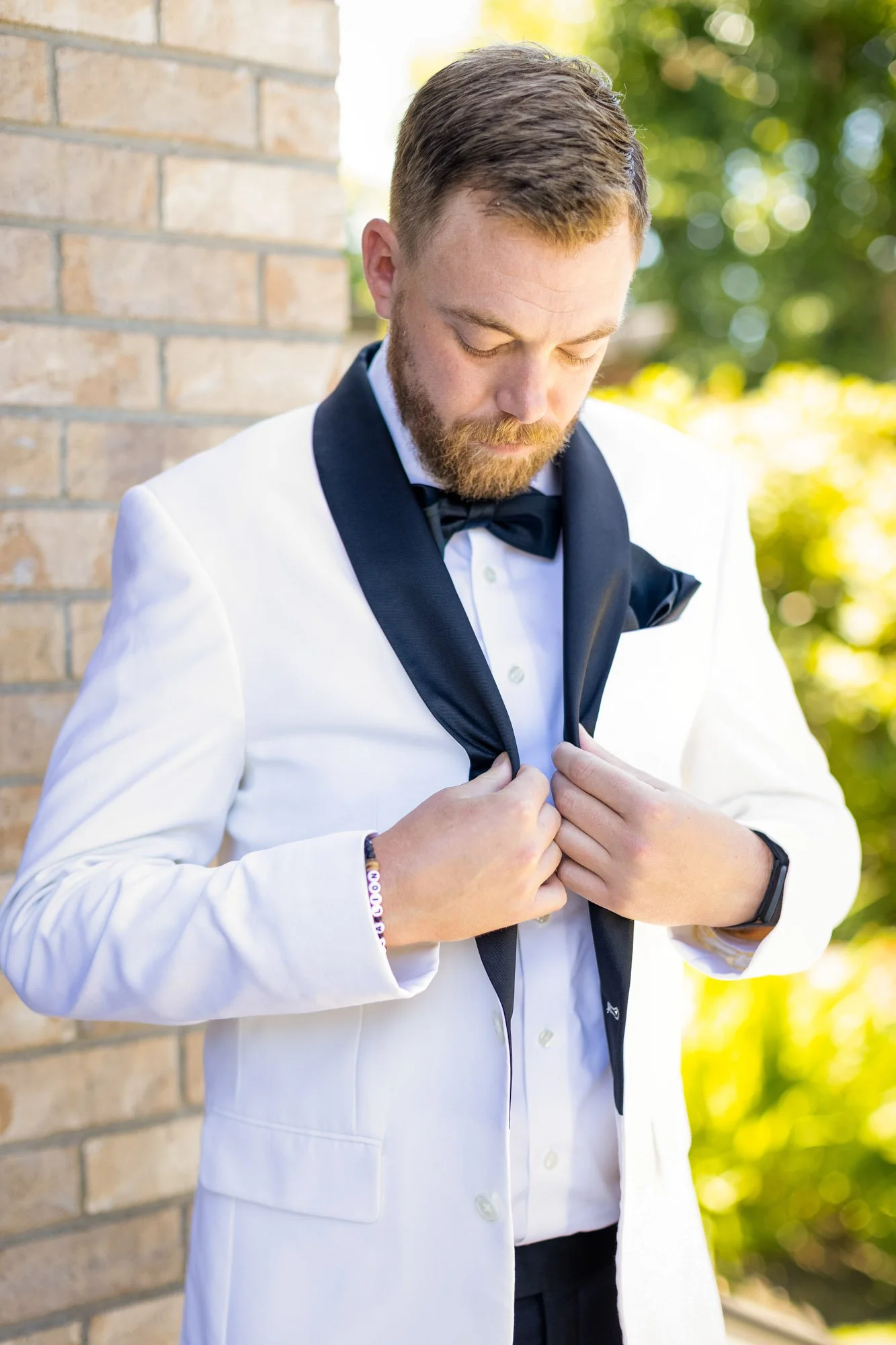Wedding photo at Abernethy Center of a groom in white tuxedo with black bow tie adjusting his tuxedo jacket outside near a brick wall, with blurred greenery in the background.