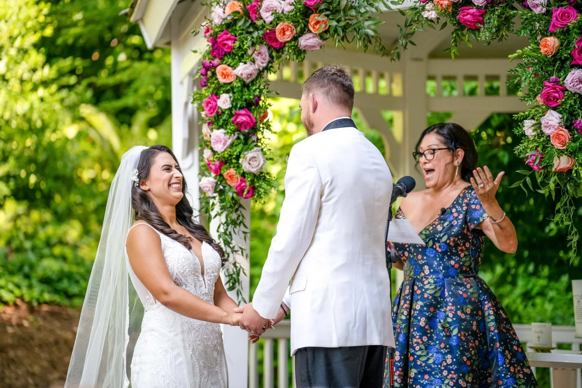 Wedding photography at Abernethy Center's Abigail's Garden of a bride and groom holding hands during a wedding ceremony outdoors, with a officiant woman speaking at a microphone.