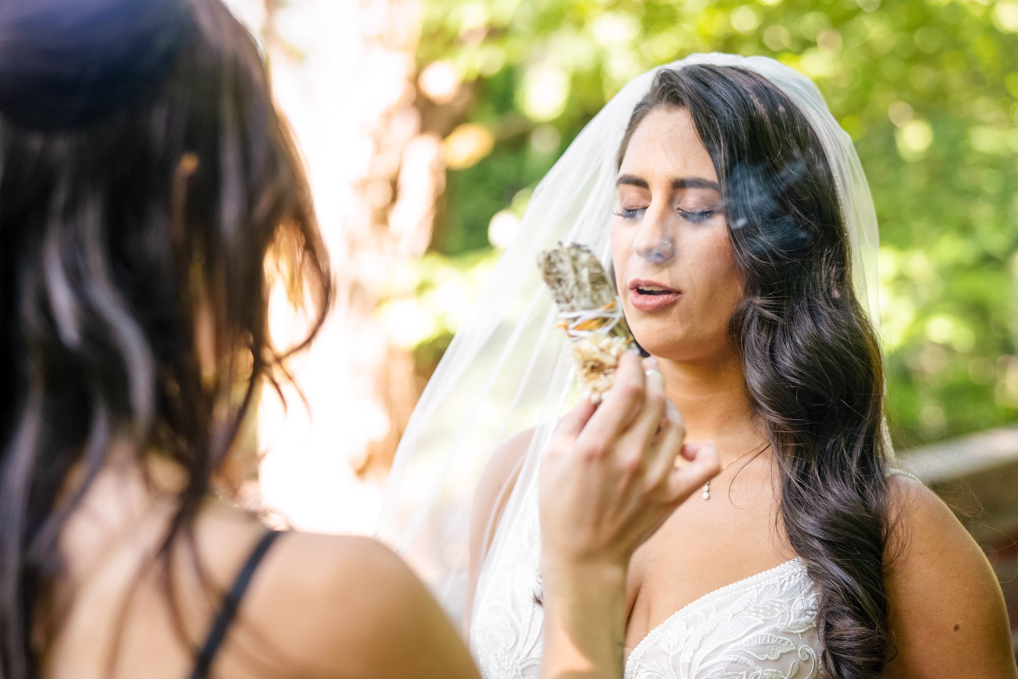 Wedding photography at Abernethy Center of A bride with dark, wavy hair and a veil is holding a decorative object near her face, with her eyes closed, in an outdoor setting with green trees in the background.