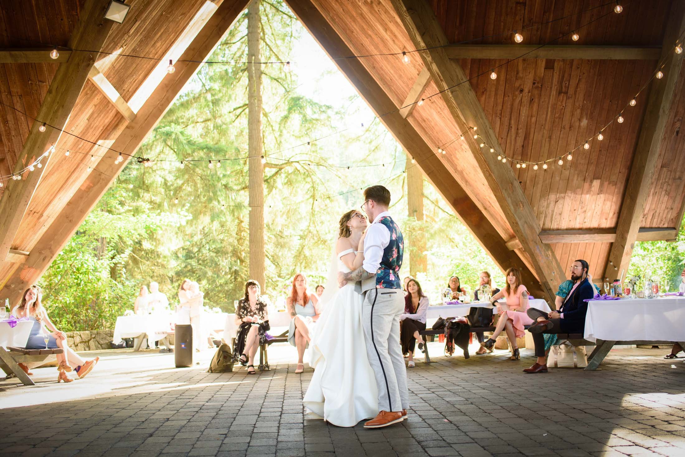 Wedding photograph at Portland's Hoyt Arboretum of A couple sharing their first dance at a wedding reception in a wooded pavilion with string lights overhead. Guests sit at tables and benches, watching the couple dance. 