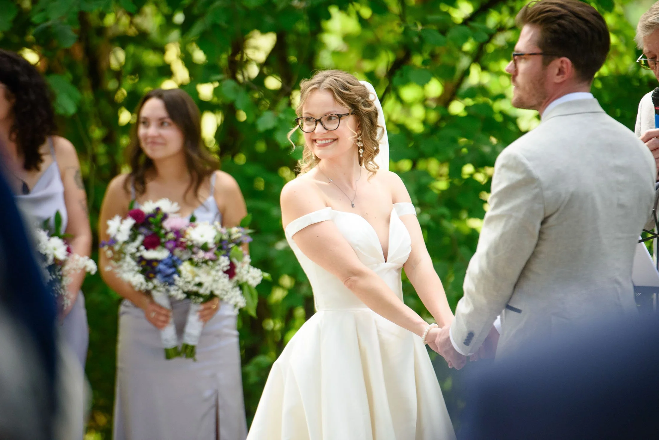Wedding photo at Hoyt Arboretum of A bride and groom holding hands during their wedding ceremony outdoors, with bridesmaids and lush greenery in the background.