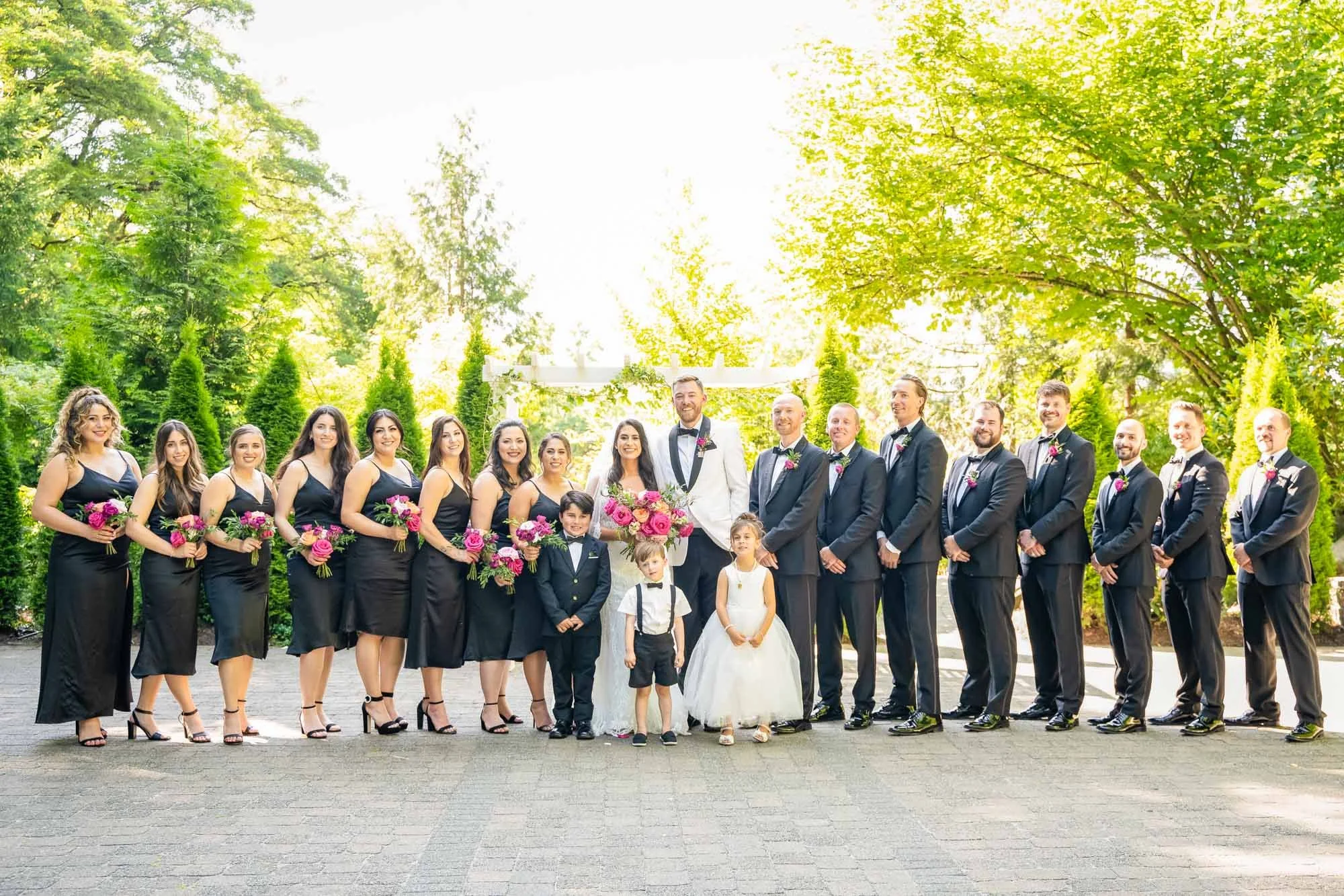 Wedding photography at Abernethy Center's Abigail's Garden of A wedding party outdoors with greenery and trees, including bridesmaids, groomsmen, a bride, a groom, and children, all dressed formally and holding bouquets.