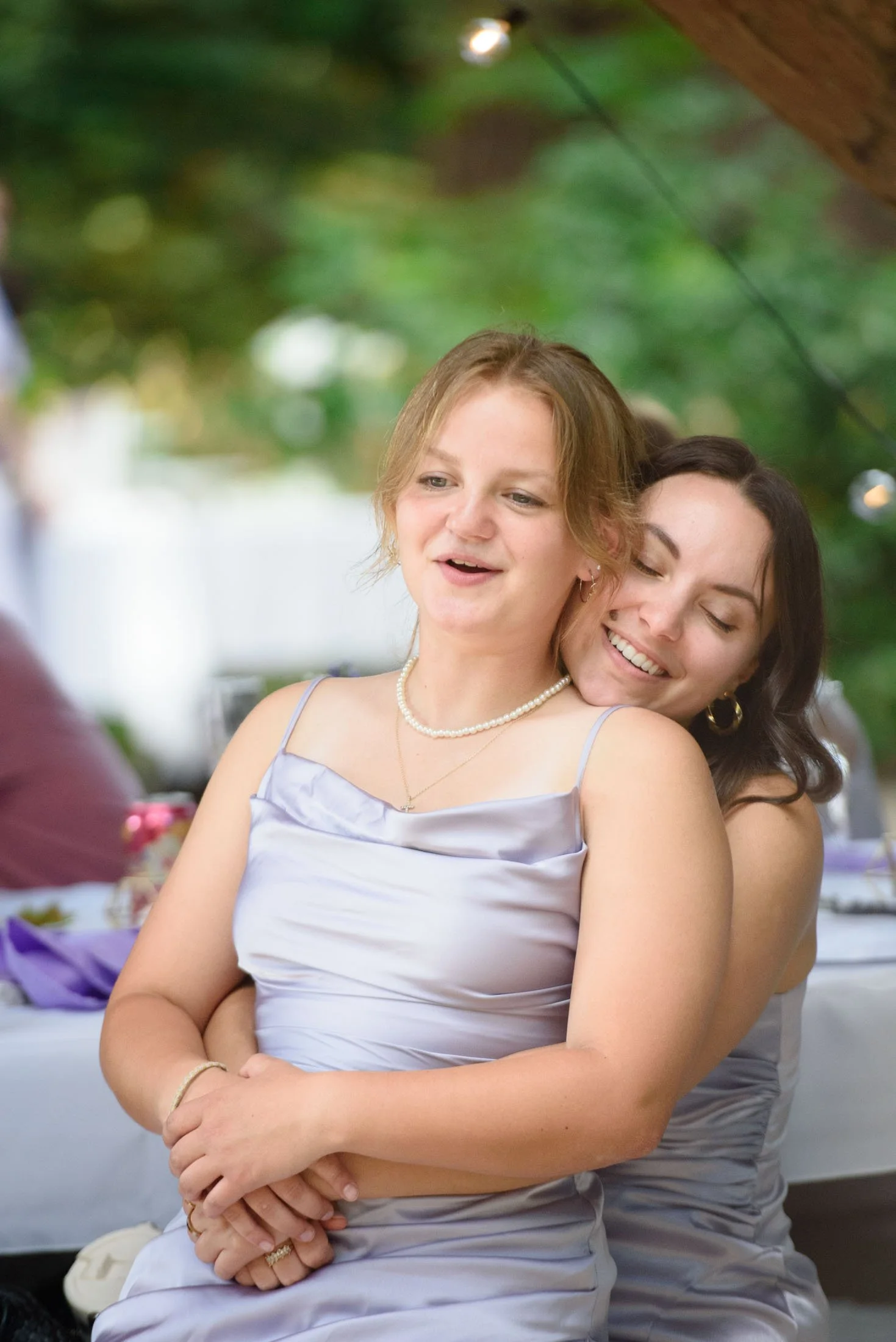Wedding photograph at Portland's Hoyt Arboretum of Two women, one with red hair and the other with dark hair, are embracing and smiling at a social gathering outdoors. They are dressed in satin dresses and appear to be happy.