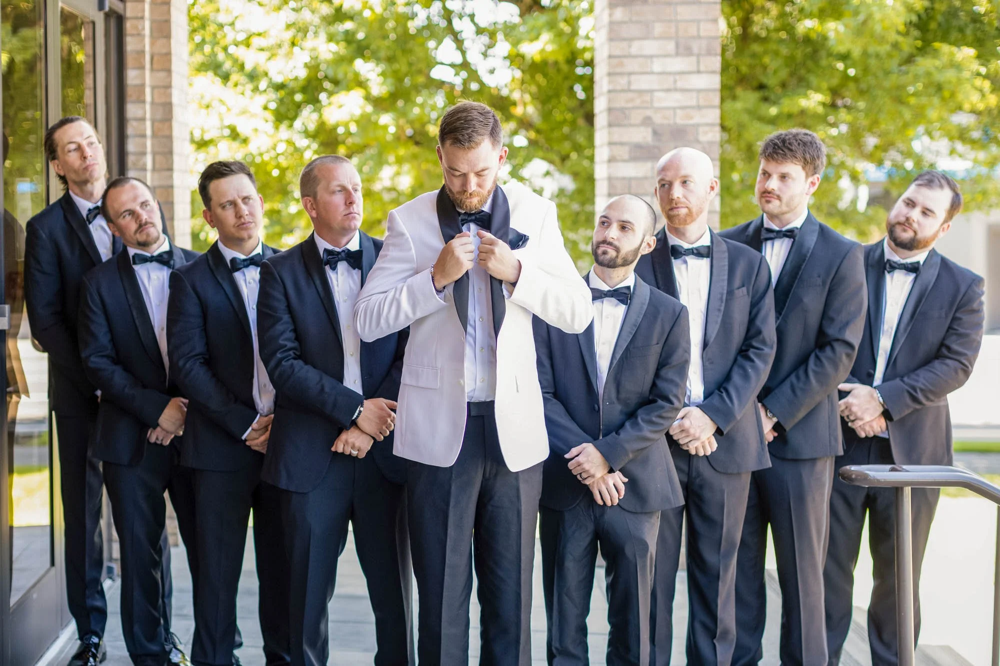 Wedding photo at Abernethy Center of A groom in a white tuxedo jacket adjusting his bow tie, surrounded by eight groomsmen in navy suits and bow ties, outside near brick and glass buildings.