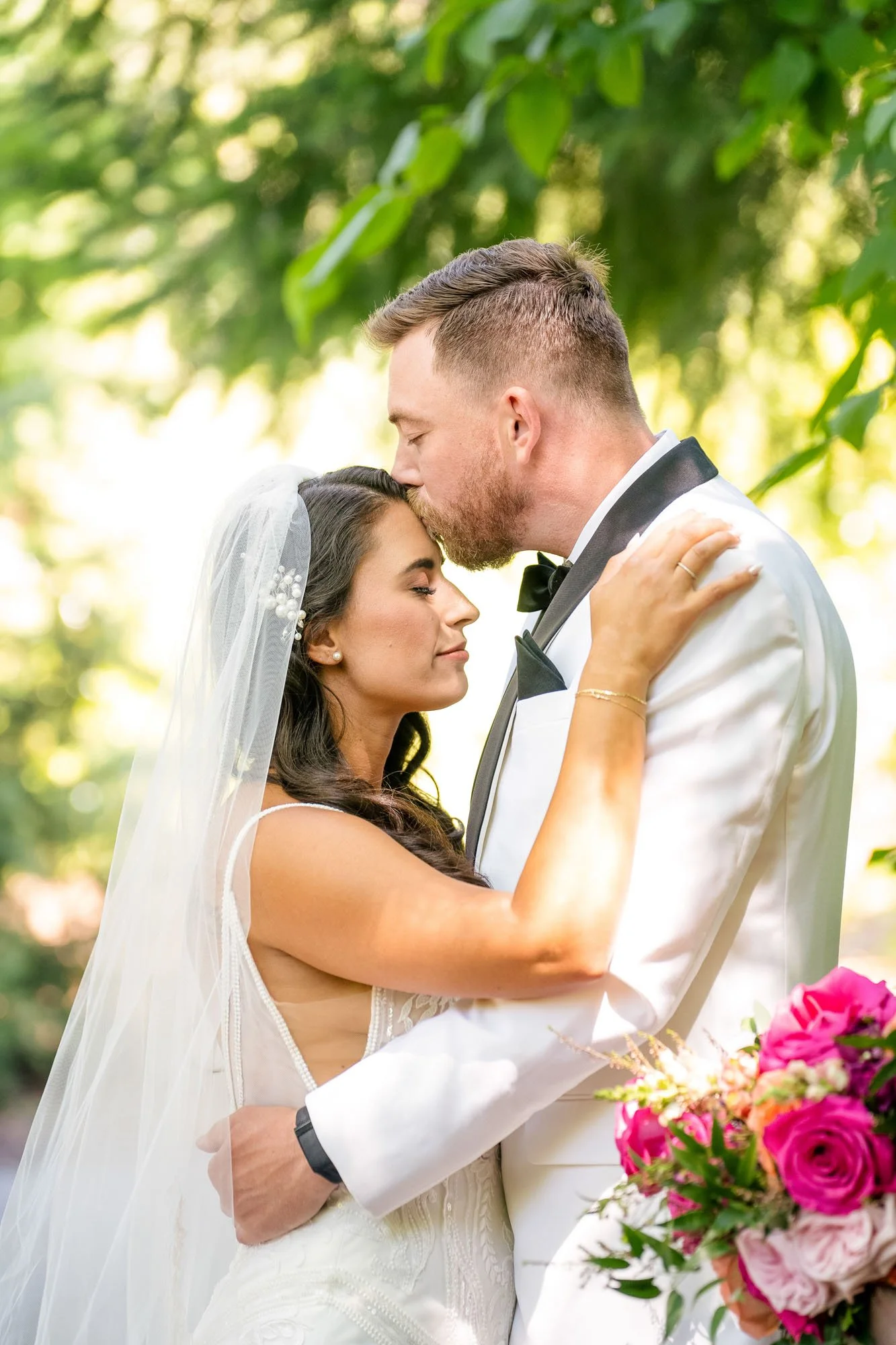 Wedding photo at Abernethy Center of A bride and groom embrace outdoors during their wedding, with the groom kissing the bride's forehead. She has dark hair, wears a veil, and holds a bouquet of pink and white flowers. 