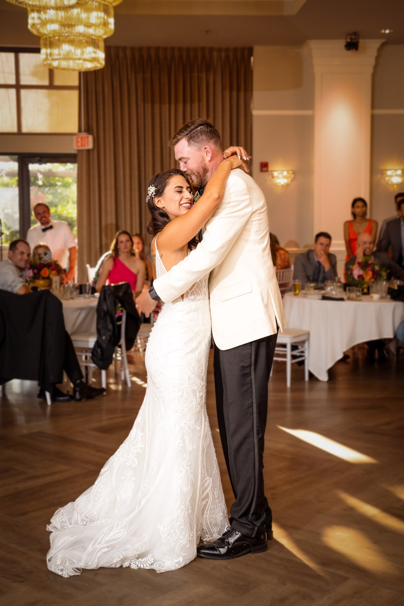 A bride and groom sharing their first dance at their wedding reception, embracing and smiling. The bride is wearing a white lace wedding dress and the groom a white tuxedo jacket with black pants. Abernethy Center Veiled Garden and Ballroom.