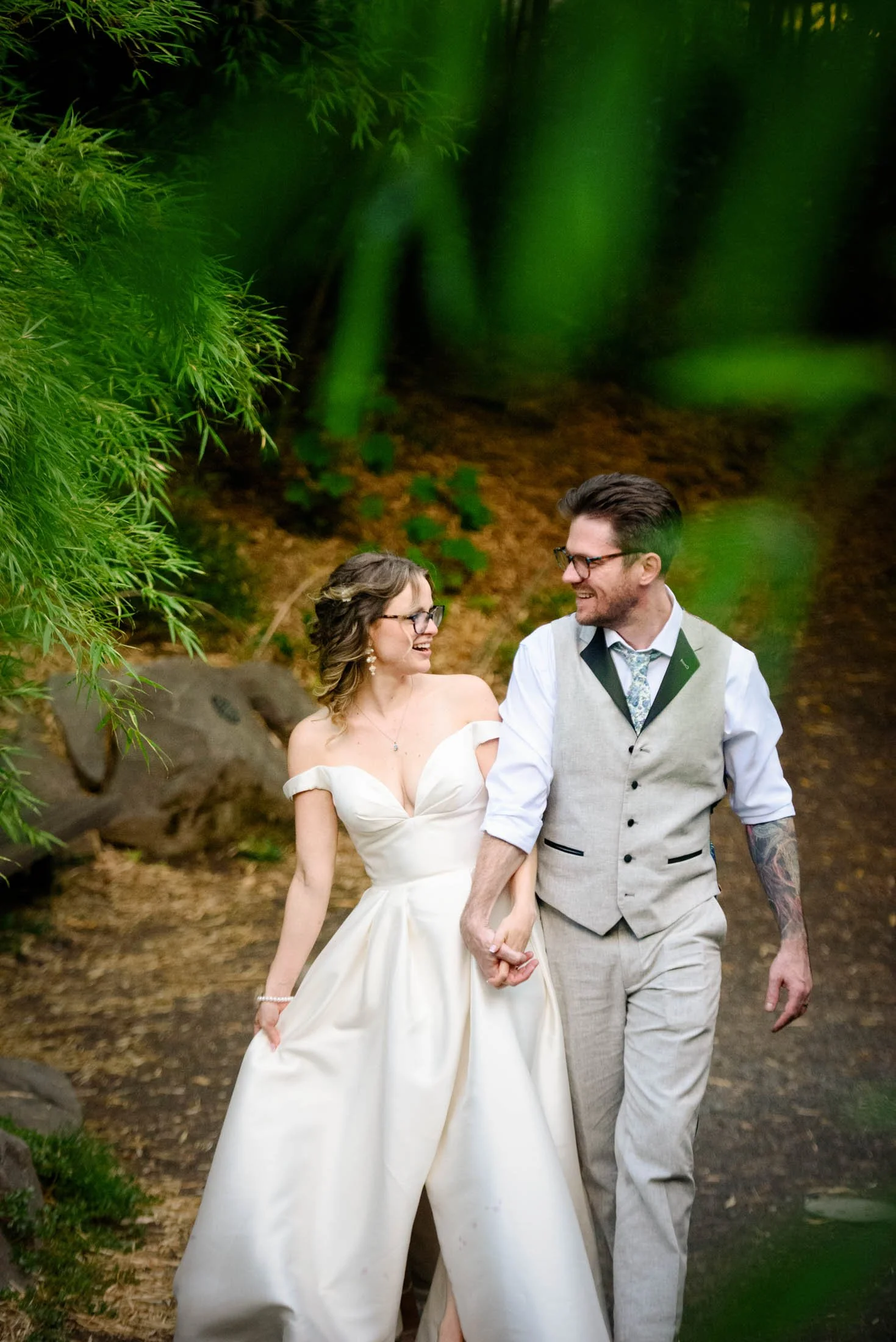 Wedding photography at Portland's Hoyt Arboretum of A bride and groom holding hands and walking in a wooded outdoor area, smiling at each other on their wedding day.