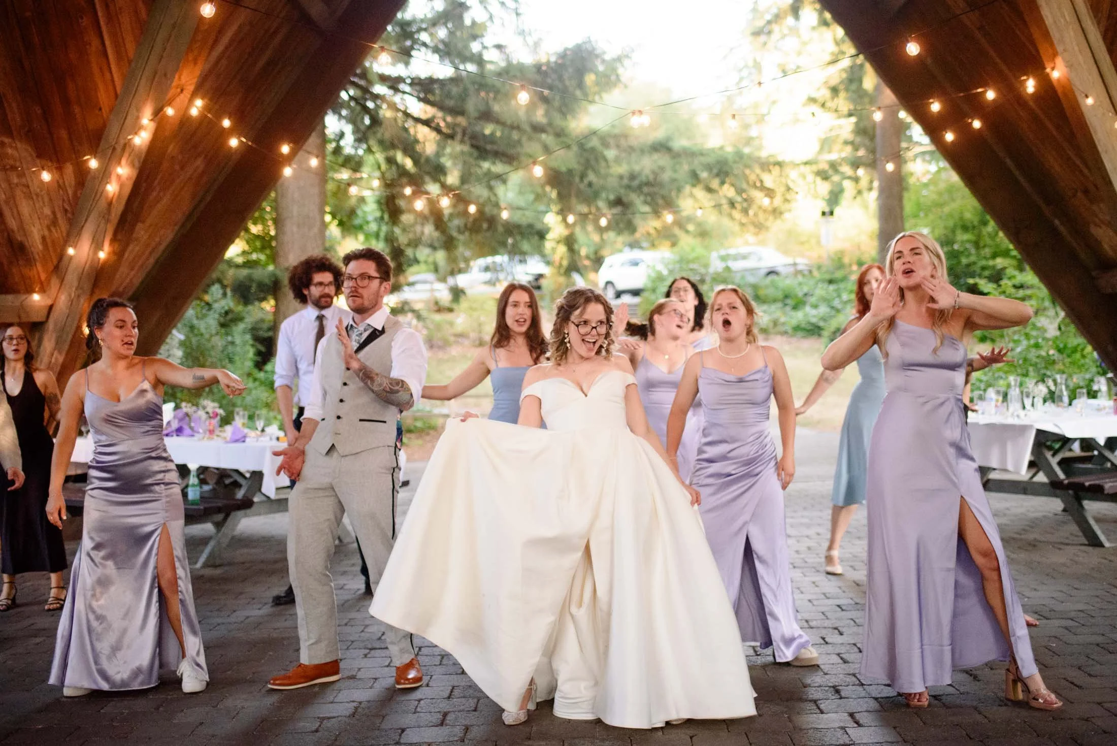 Wedding photograph at Portland's Hoyt Arboretum of A bride in a white gown dances with bridesmaids and guests under a wooden pavilion decorated with string lights during a wedding reception.