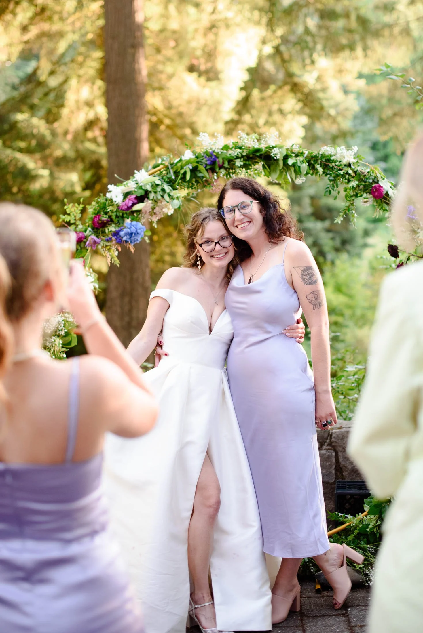 Wedding photograph at Portland's Hoyt Arboretum of two women standing under a floral arch at an outdoor wedding, smiling and posing for a photo. One is in a white wedding dress and the other in a lavender dress, with trees and sunlight.