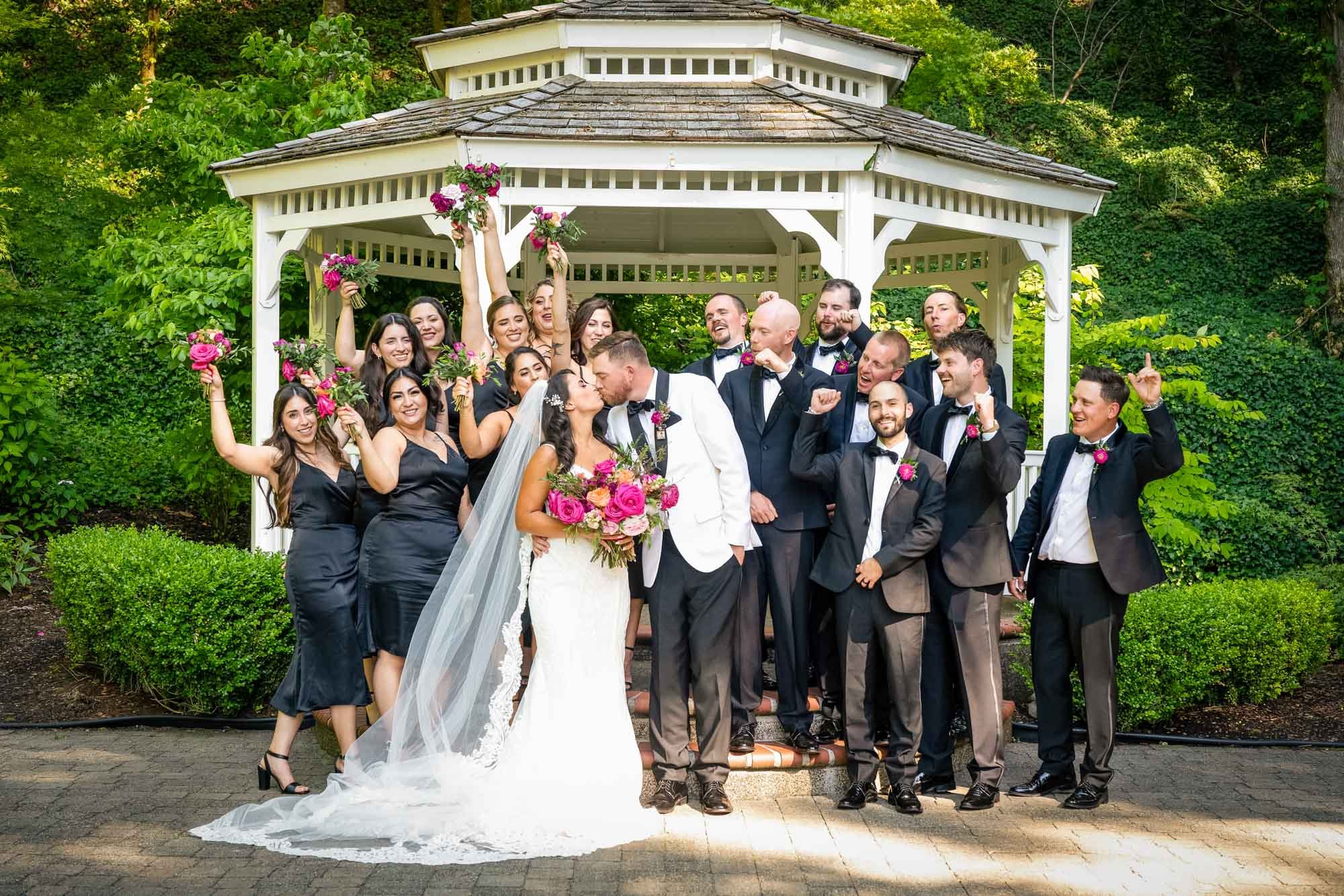 Wedding photo at Abernethy Center's Abigail's Garden of wedding party of 20 people, including a bride and groom, celebrating in front of a gazebo surrounded by green trees. The bride is in a white wedding gown holding a bouquet of pink flowers.