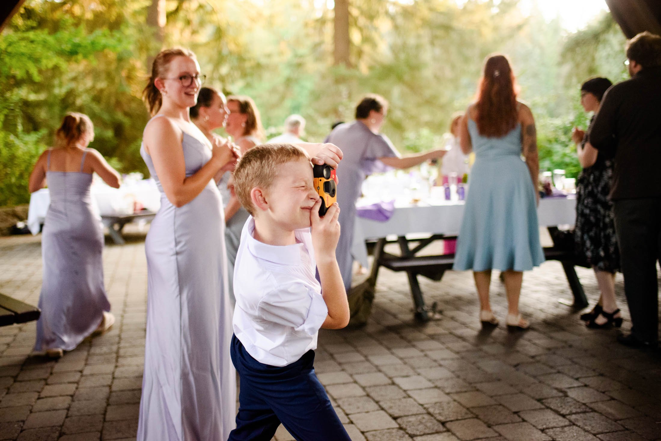 A young boy with a camera in his hand takes a picture at a gathering of people outdoors on a paved patio surrounded by trees. The background shows women in dresses near a table with food and drinks.