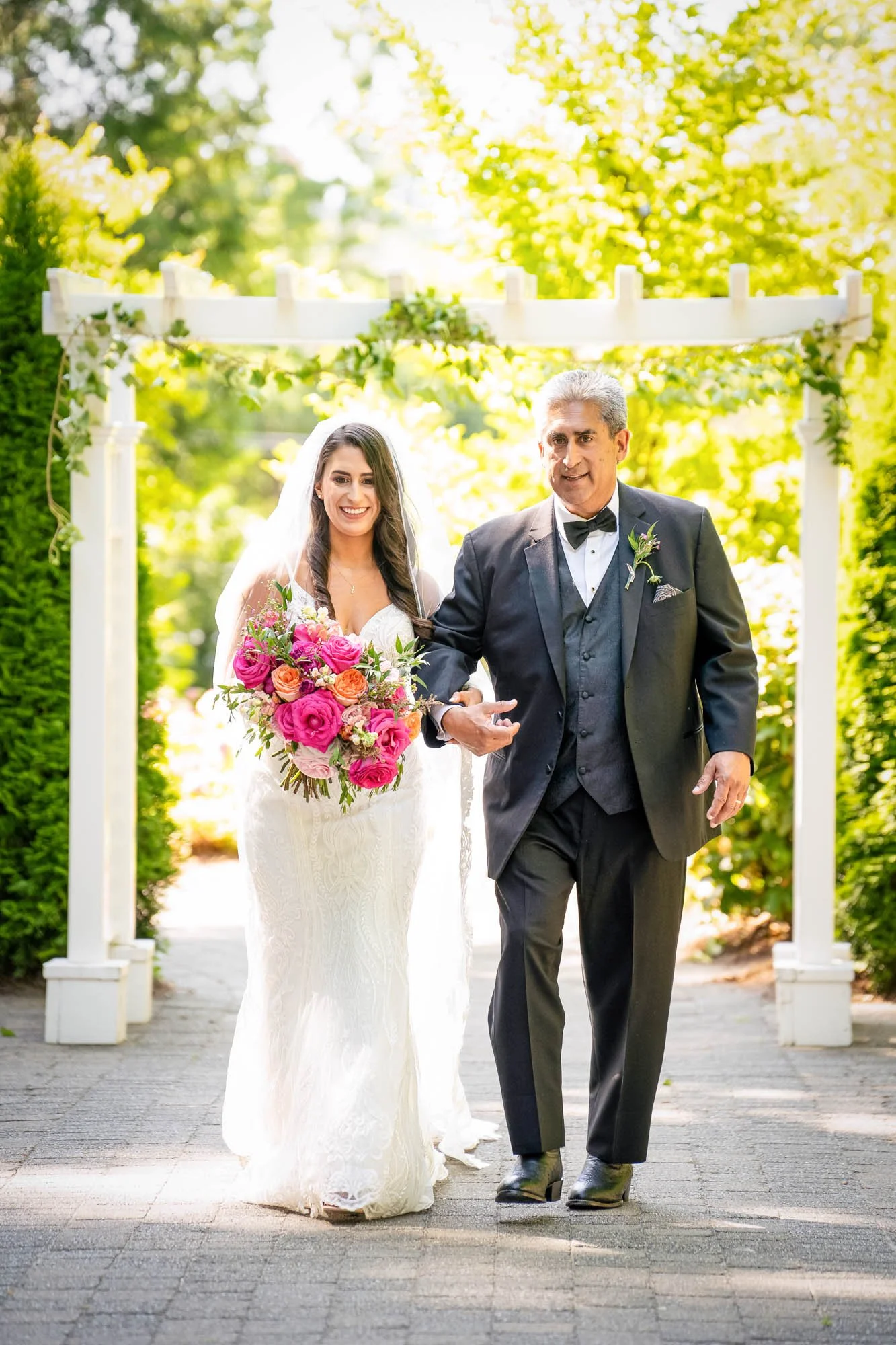 Wedding photography at Abernethy Center of a Bride in a white wedding dress holding a bouquet walking with an older man in a tuxedo under a white arbor outdoors surrounded by green trees.