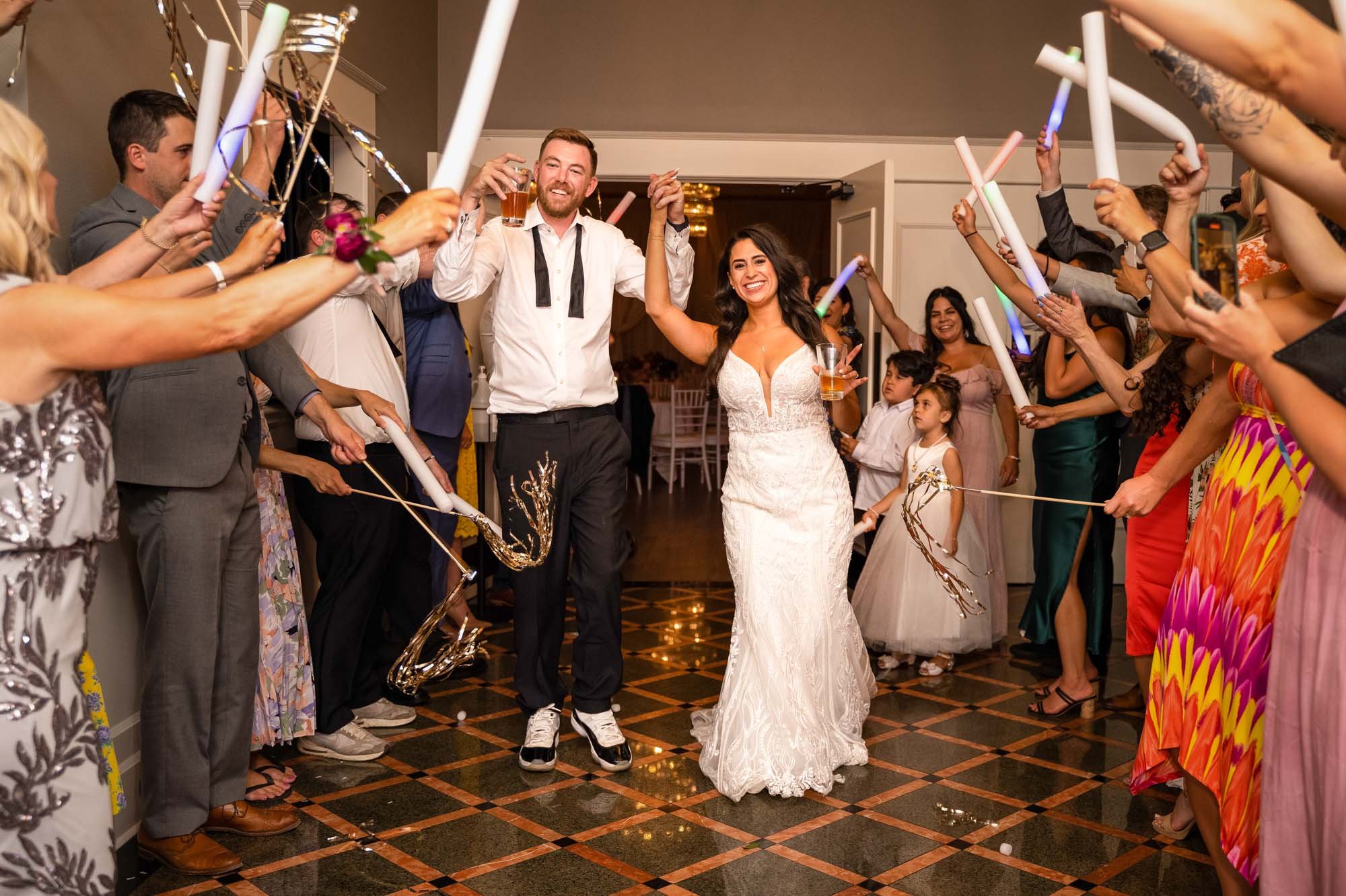 Wedding photograph at Abernethy Center Veiled Garden and Ballroom of A newlywed couple celebrating their wedding with guests holding glow sticks and cameras, walking down a decorated hall.