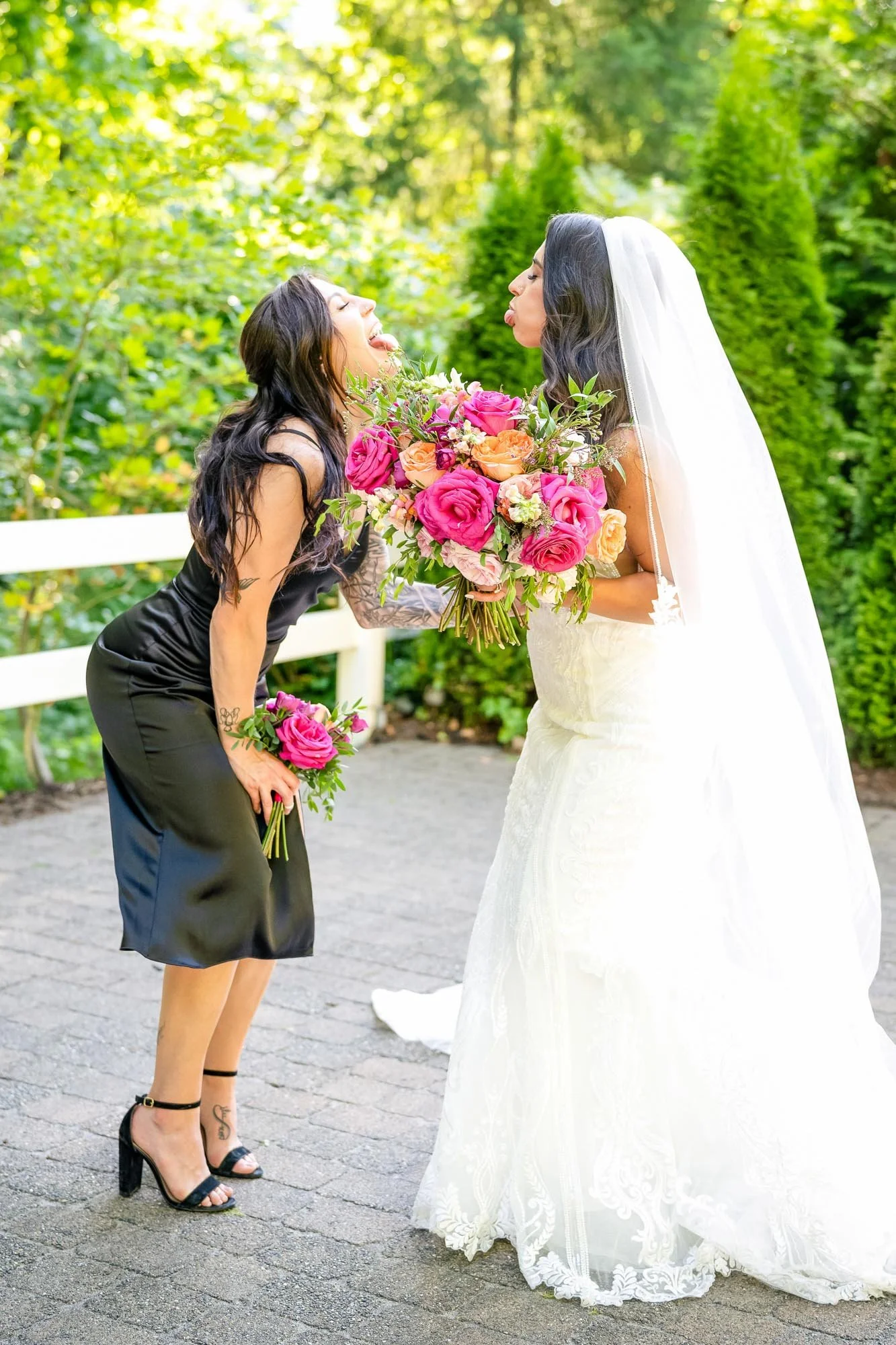 Wedding photo at Abernethy Center's Abigail's Garden oTwo women, one in a wedding dress and the other in a black dress, are outdoors with greenery in the background. They are holding pink and peach bouquets and are making playful faces at each other.