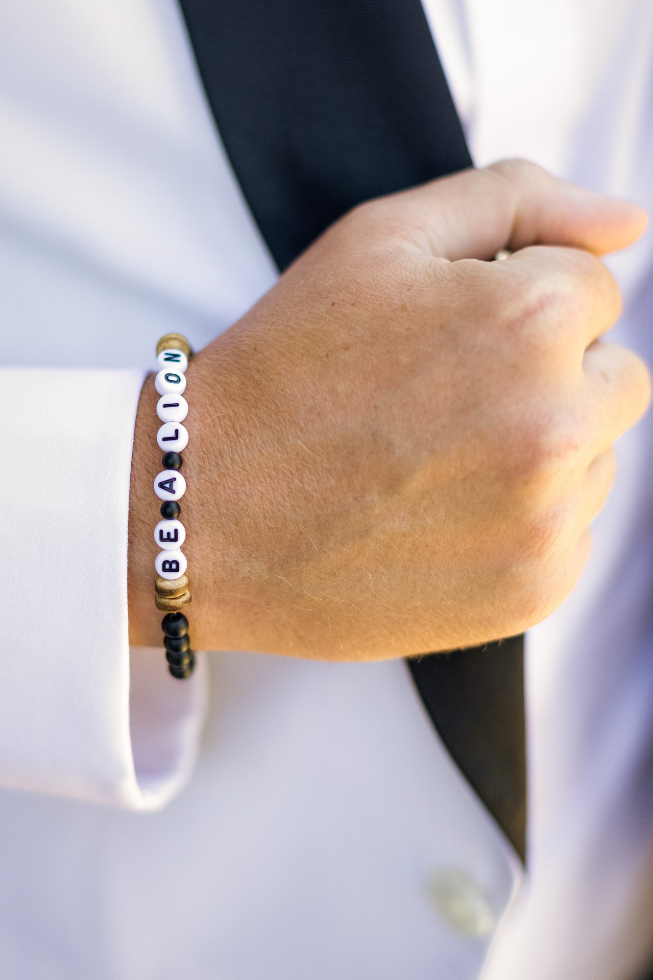Close-up of a person's wrist wearing a beaded bracelet that spells out 'BEA LOV N' and part of their hand, with a white shirt and black tie.