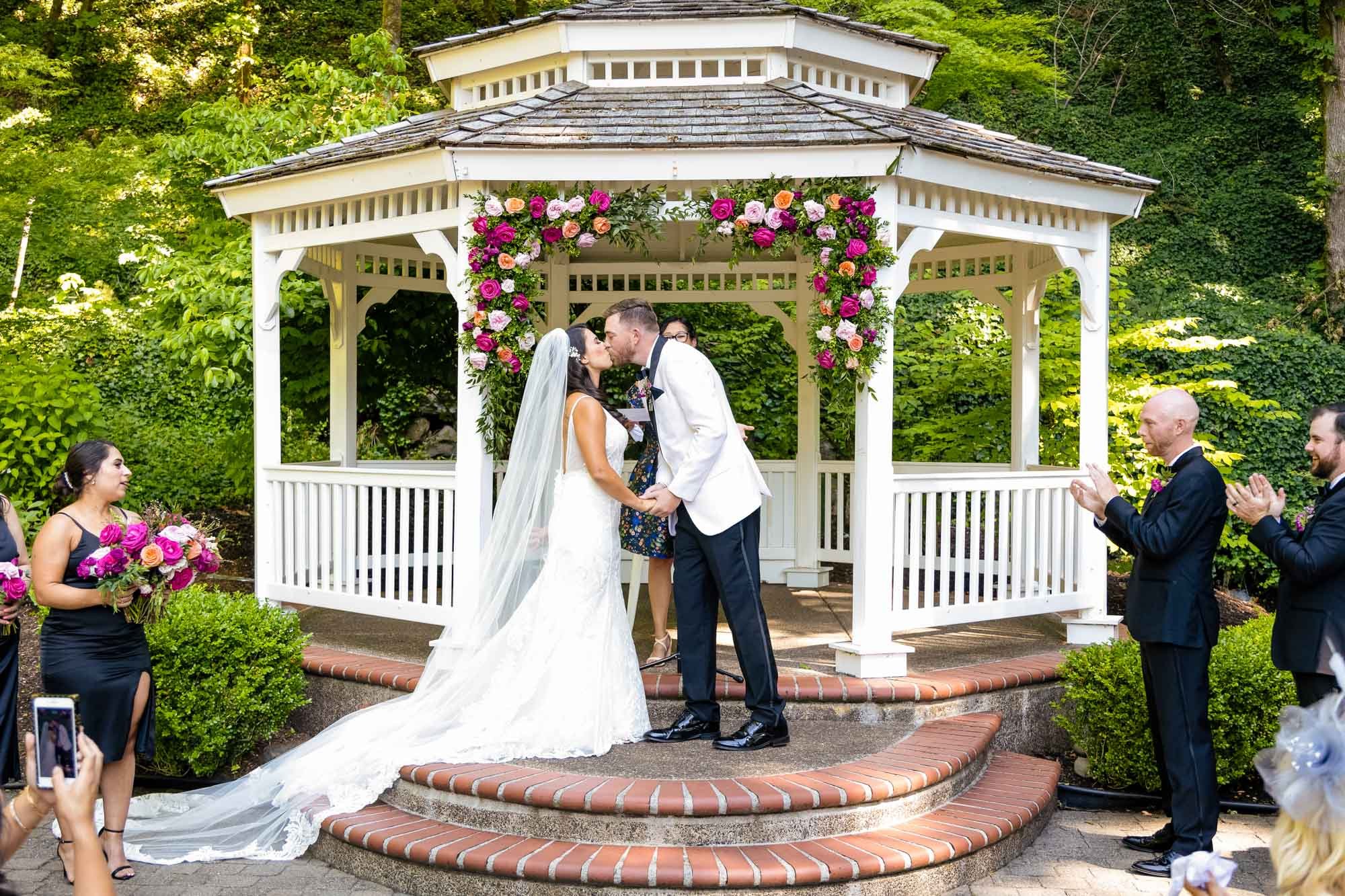 Wedding photography at Abernethy Center's Abigail's Garden taking place outdoors under a white gazebo decorated with pink and purple flowers. The bride and groom are kissing, holding hands, with the bride wearing a white wedding gown and veil.