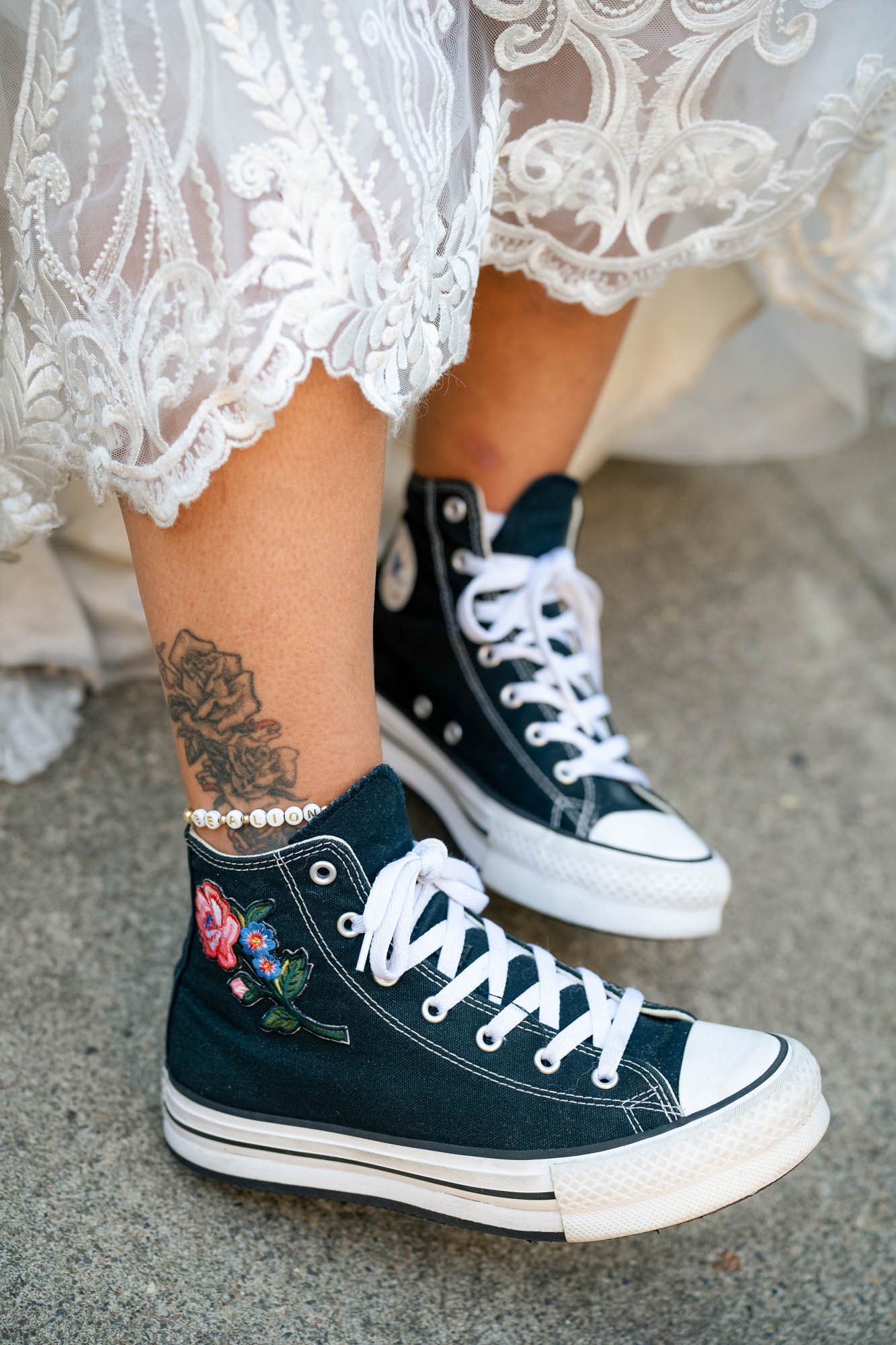 Person wearing black high-top Converse sneakers with floral embroidery, lace-trimmed white lace dress, and a tattooed ankle with a beaded bracelet.