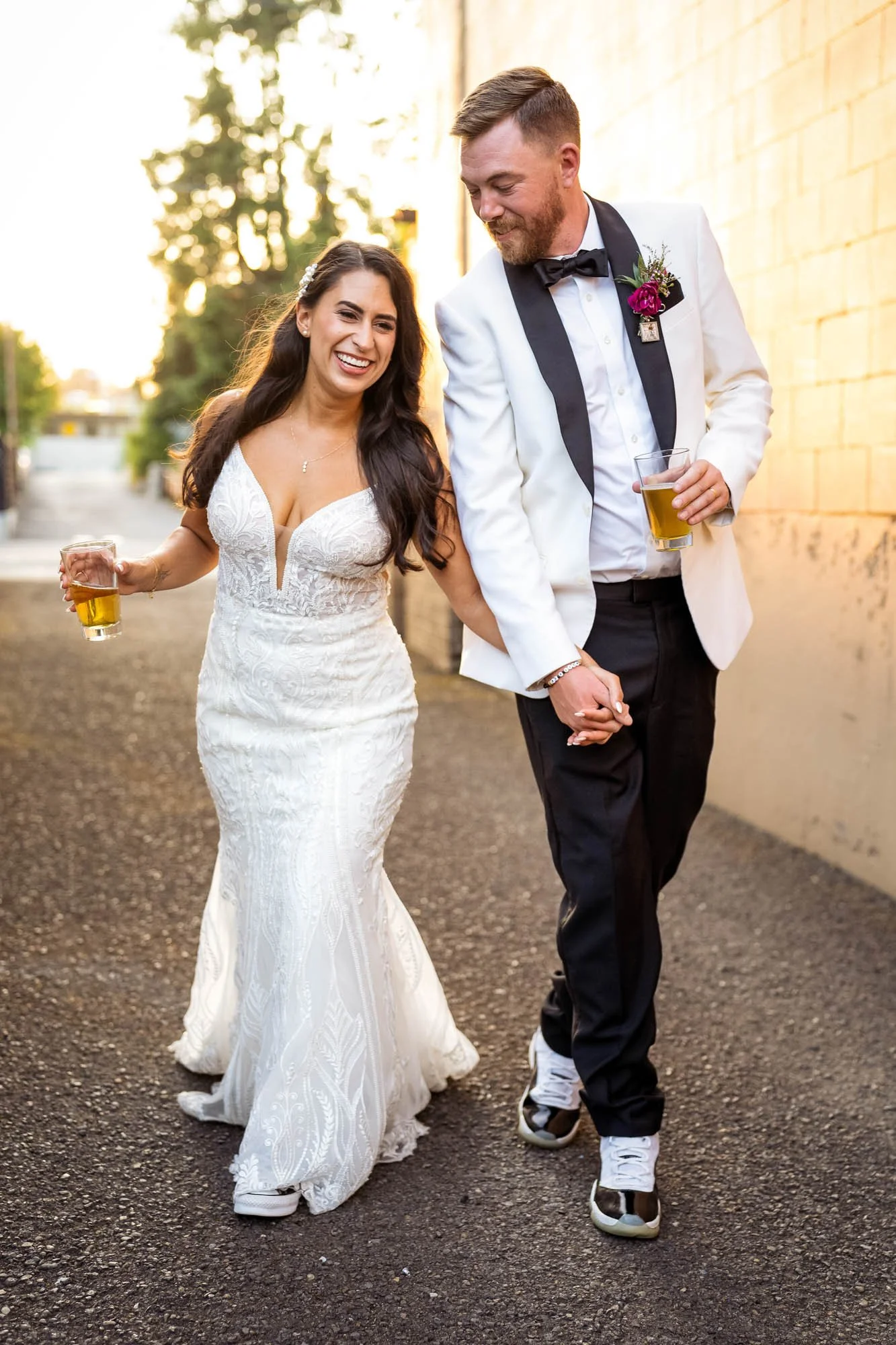 A newly married couple in wedding attire walking outdoors, holding hands and glasses of beer, smiling at each other in warm sunlight at Abernethy Center Veiled Garden and Ballroom.
