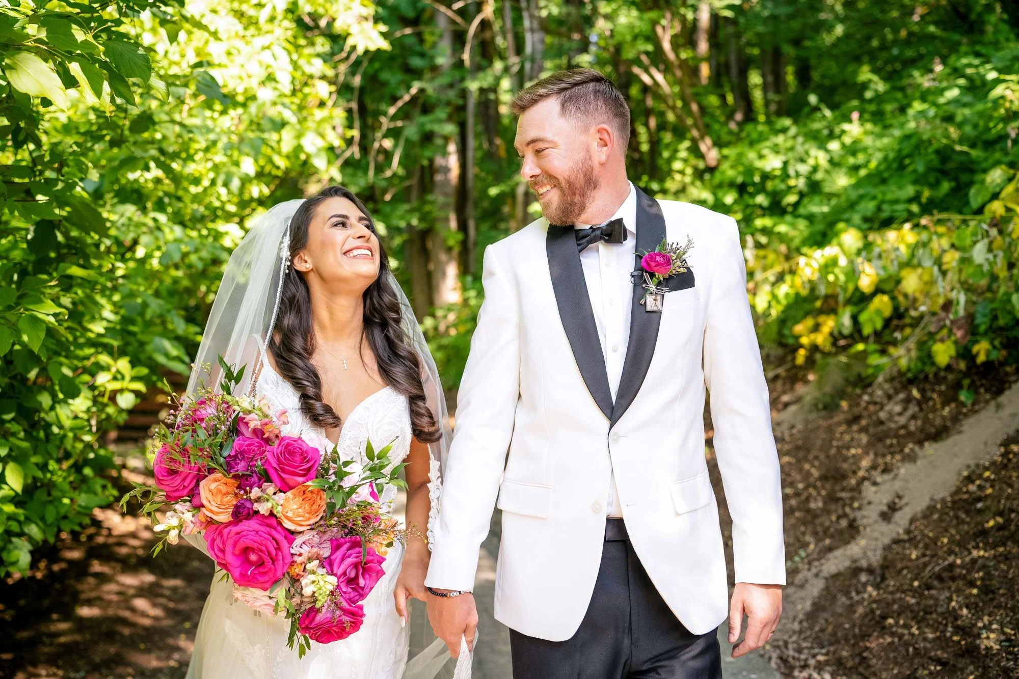 A bride and groom holding hands and walking in a lush green outdoor setting, smiling at each other. The bride is wearing a white wedding dress and veil, holding a bouquet of pink and orange flowers. The groom is in a white tuxedo jacket with black la