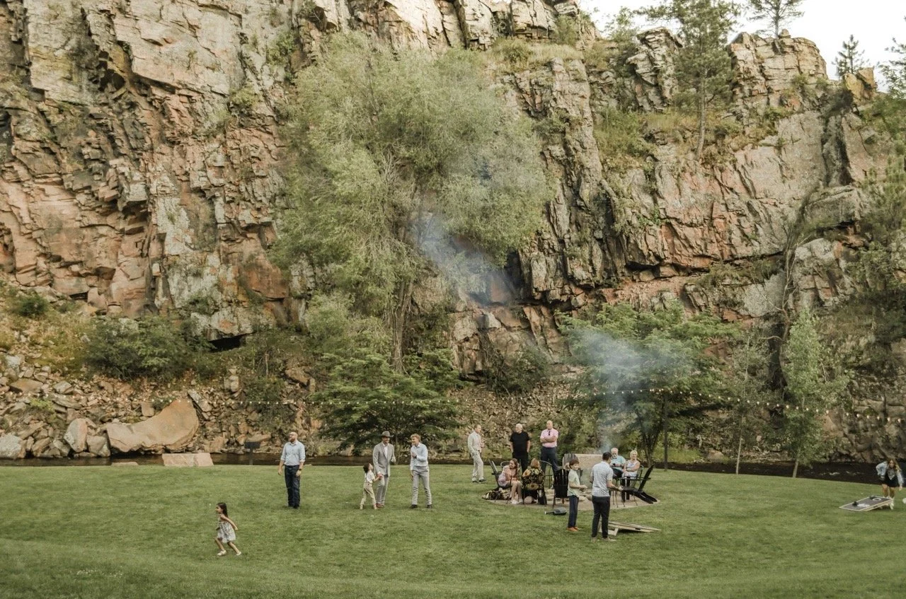 Guests sitting by a fire outdoors, surrounded by trees and rocks, sharing a romantic moment in a natural setting.
