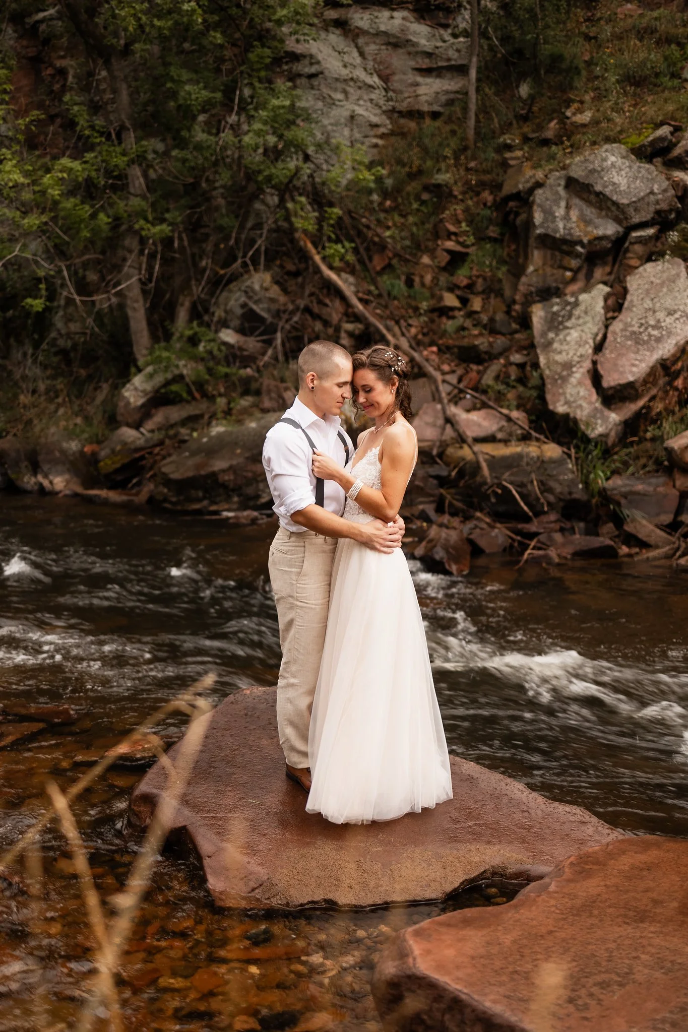 A couple dressed in wedding attire stands close together on a large flat rock in a river, surrounded by rocks, water, and trees in a natural outdoor setting.