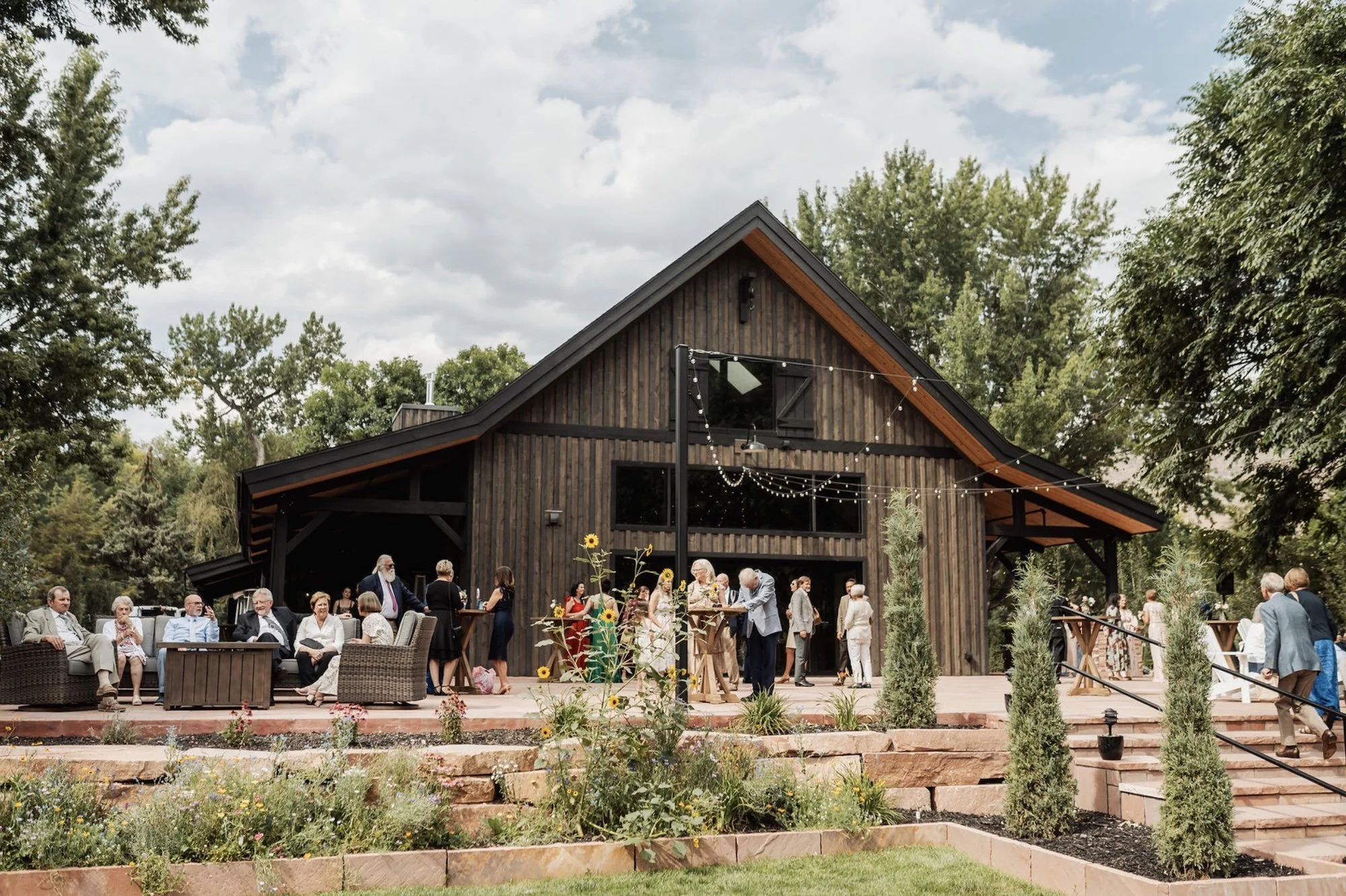 People gathered outside a modern rustic wooden barn decorated for a celebration with string lights and flowers.