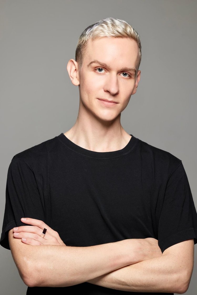 Young man with short blonde hair, light skin, wearing a black T-shirt, standing against a plain gray background.