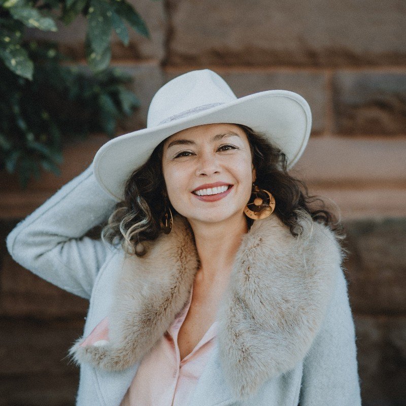 Woman smiling outdoors, wearing a white hat, a fur-collared coat, and large gold earrings.