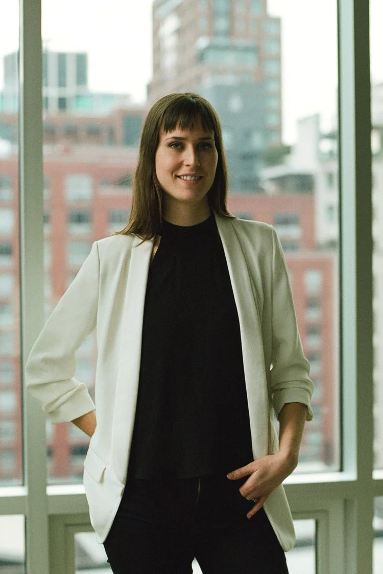 A woman standing in front of large windows with a cityscape background, wearing a white blazer over a black top.