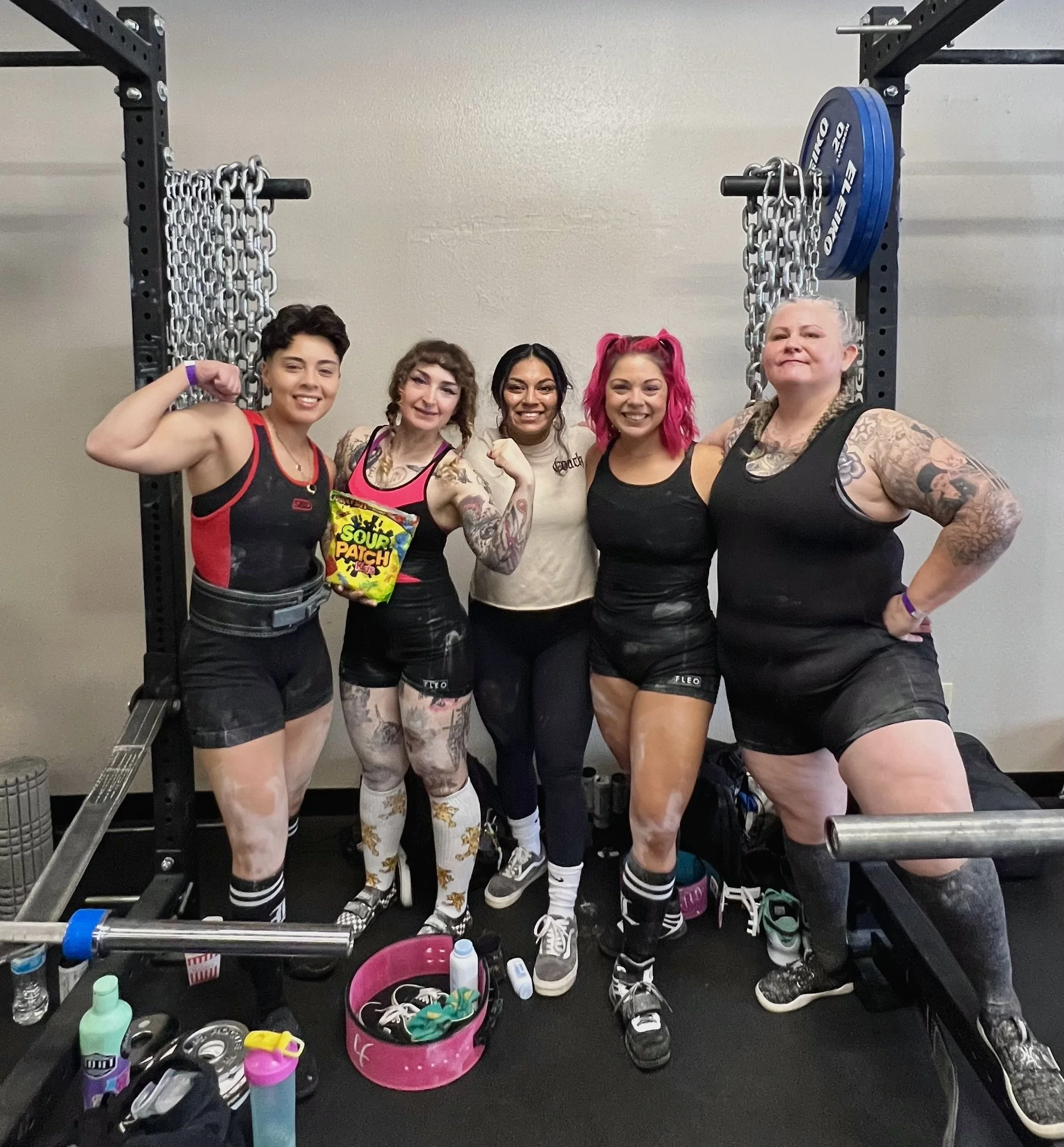 Group of five women posing together in a gym, some flexing muscles, in workout attire, with gym equipment and supplies around.