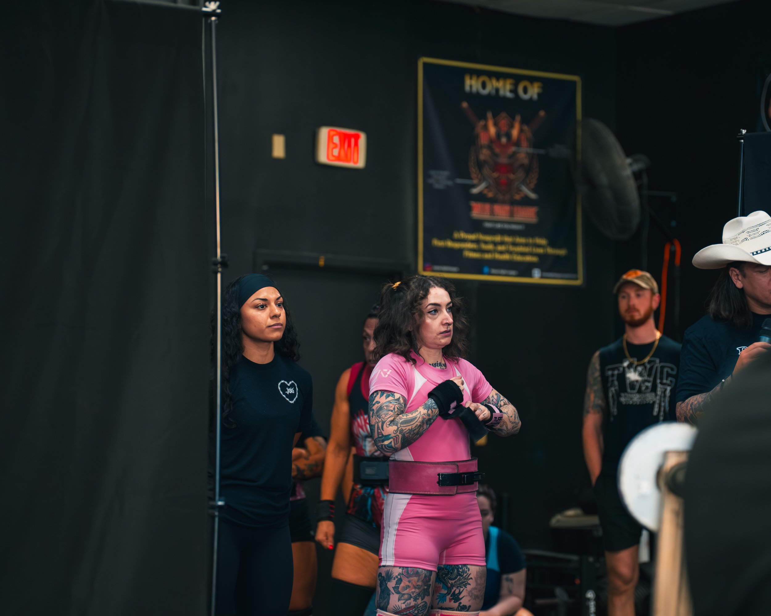 Women at a fitness event, one in a pink workout outfit with tattoos, enlisting in a gym or training facility with others in background, dark interior, sign on wall, and gym equipment.