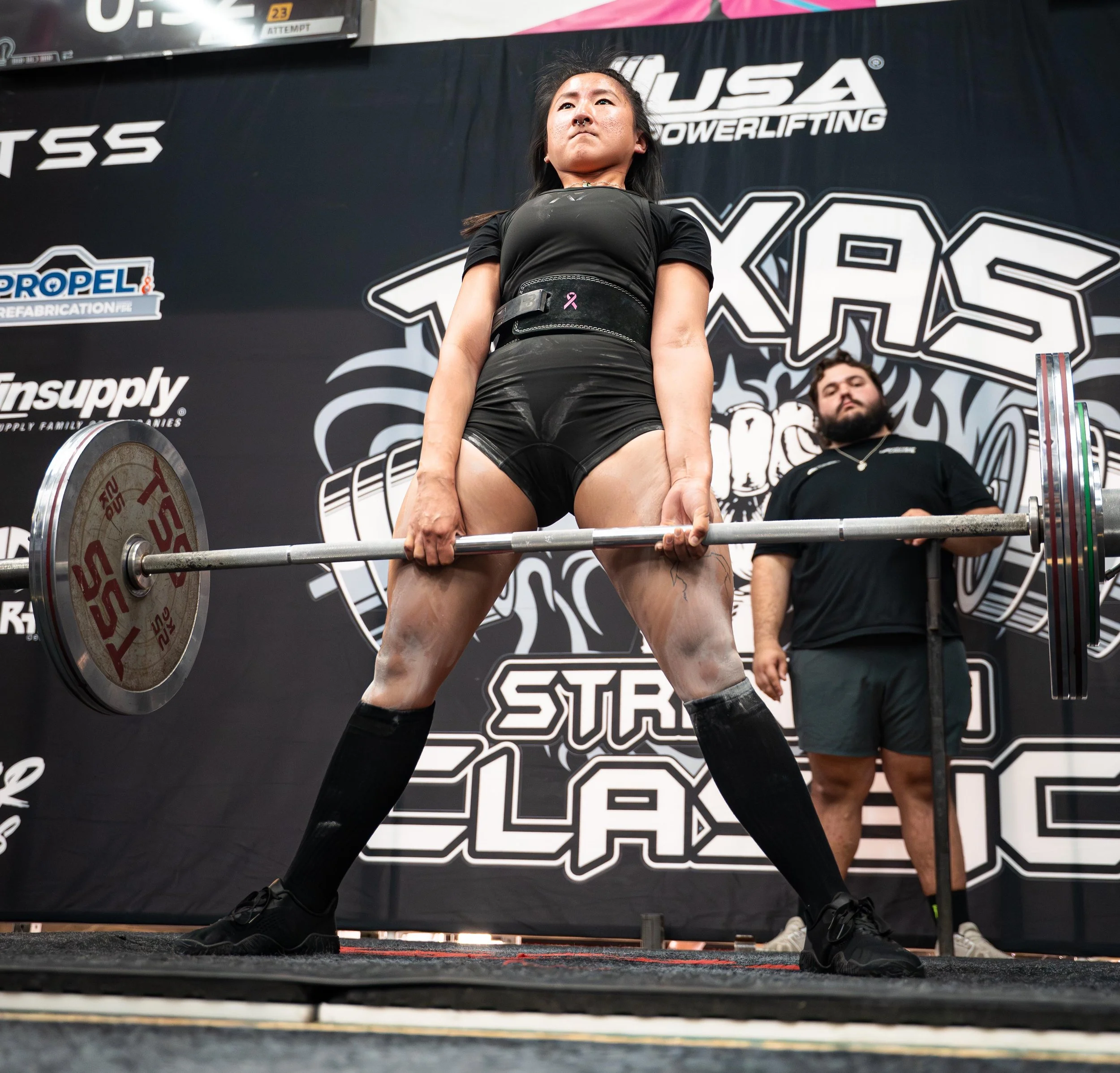 A female powerlifter in black attire is performing a deadlift, holding a loaded barbell on a stage at a competition. She is wearing a lifting belt and knee-high socks, with a male spotter or coach standing behind her observing.