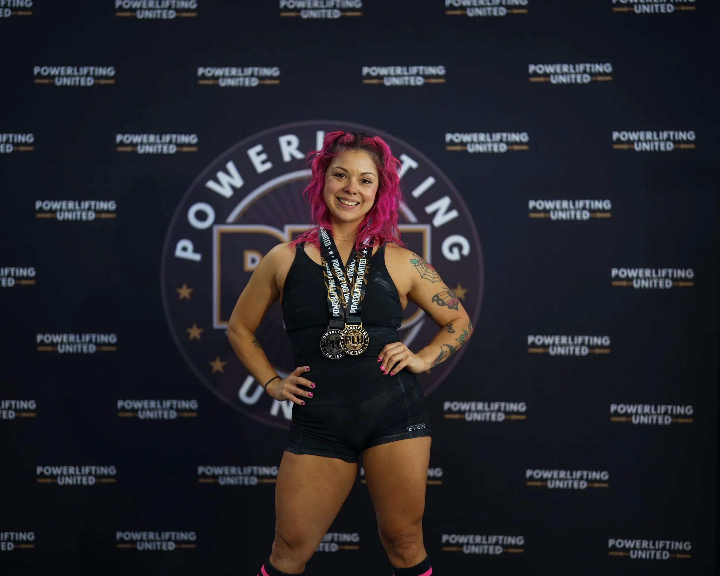A woman with pink hair smiling and wearing a black sports outfit, standing in front of a backdrop with 'Powerlifting United' logos and a medals around her neck.